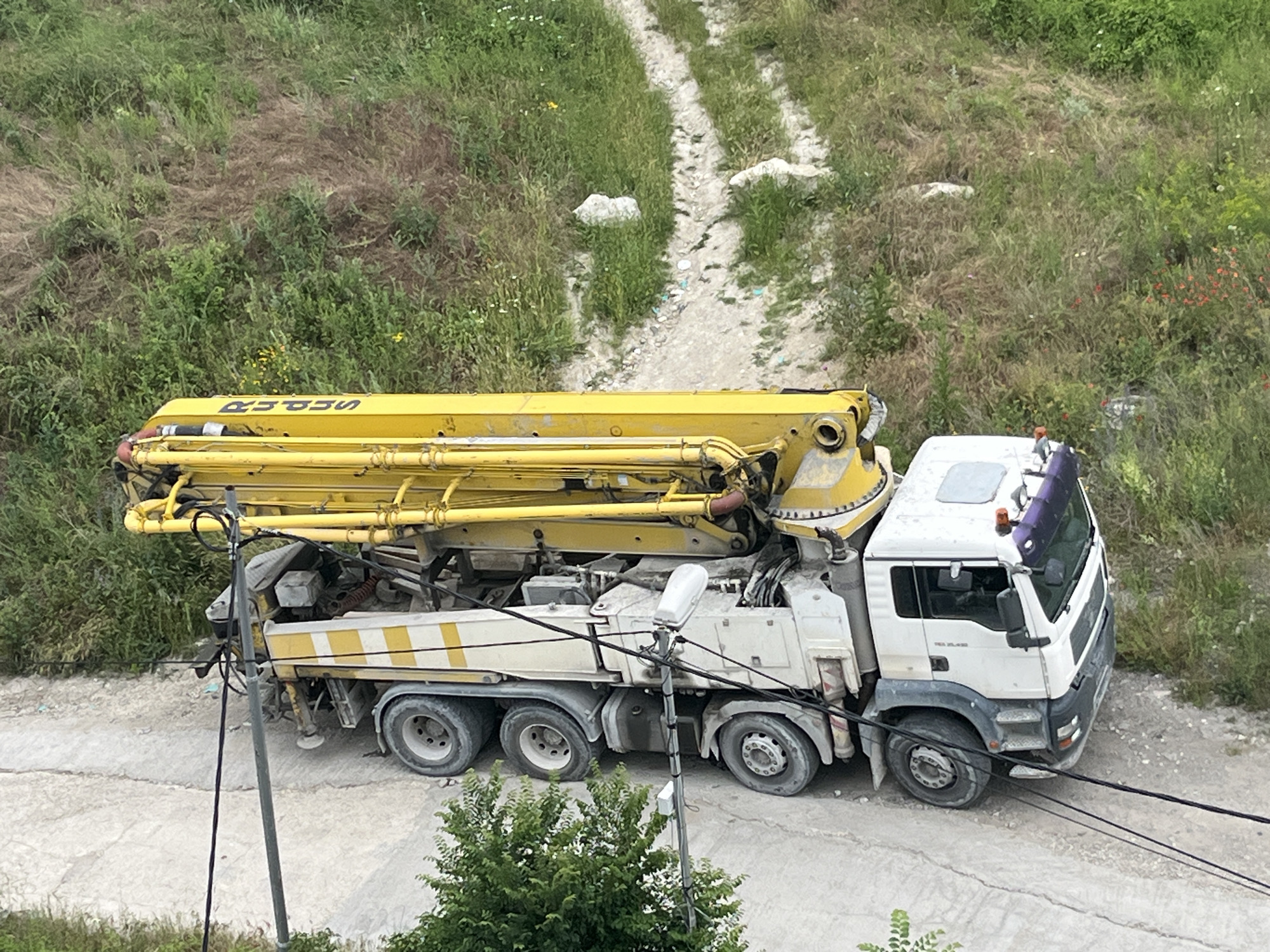 In Varna, Bulgaria, a concrete pump truck, predominantly yellow and white, is parked on a dirt and concrete surface. The truck features a long, extendable yellow boom. The surrounding environment is overgrown with green vegetation, including grass, weeds, and some wildflowers. A dirt path with tire tracks runs through the grassy hillside behind the truck. The scene is captured from a slightly elevated, aerial perspective. No people are visible in the image.