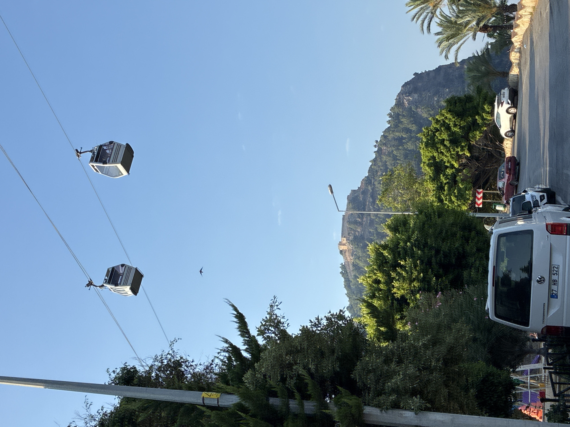 The image captures a view of Alanya, Turkey, with two cable car cabins suspended over a clear blue sky. In the foreground, a grey utility van with license plate "27 HB 522" is parked on a paved road. A red sports car and a white SUV are also visible on the road, which curves to the right. Palm trees and lush green trees line the roadside, leading to a rocky mountainside. Atop the mountain, remnants of a historical fortress are visible. A lamppost stands near the mountain's base. A utility pole with a yellow warning sticker is on the left side of the frame, partially obscured by dense evergreen foliage. A small bird is captured in flight in the upper portion of the sky.
