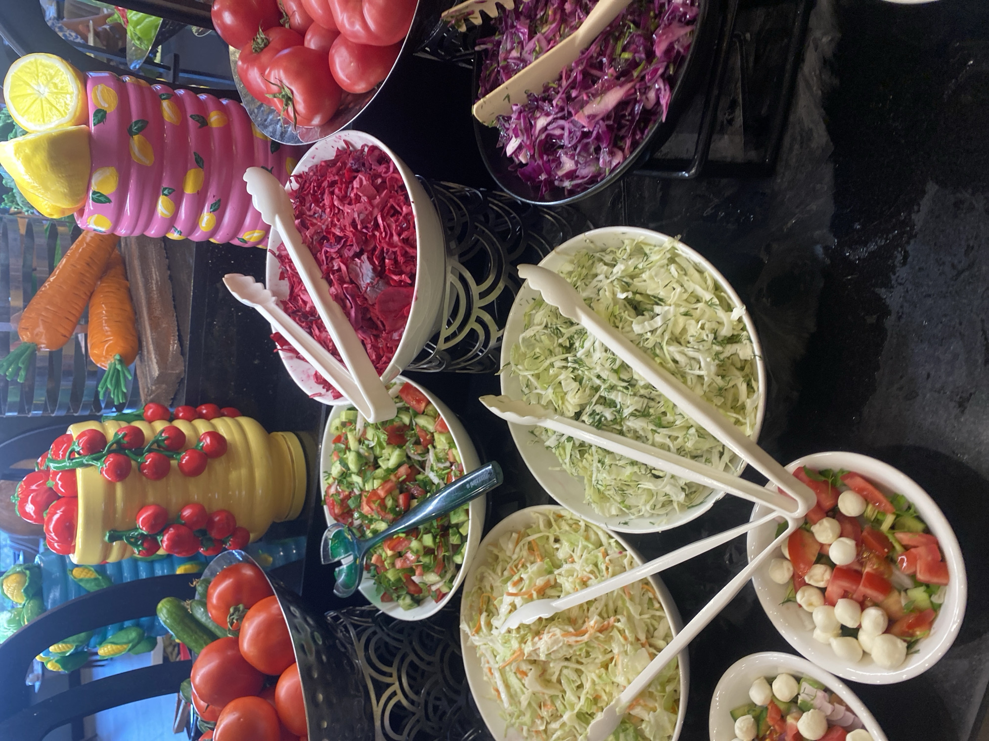 The image displays a buffet selection of fresh salads and tomatoes. In the foreground, a white oval bowl contains a salad of chopped tomatoes, small white mozzarella balls, and herbs, with white tongs reaching into it. To its left is another white oval bowl with a similar salad. Behind these, several other bowls are visible: one with shredded cabbage and carrots in a creamy dressing, another with finely shredded green cabbage and dill, and a larger bowl with shredded red cabbage. A bowl of chopped red cabbage and another of chopped tomatoes, cucumbers, and herbs are also present. In the background, a metal stand holds a bowl of shredded red vegetables, possibly beets. Various whole tomatoes are displayed in a metal bowl. Decorative items, including a pink vase with lemon motifs and a stack of orange carrot-like objects, are positioned behind the food. The buffet is set against a black, reflective surface. No people are visible in the image. The location is specified as Aksu, Turkey.