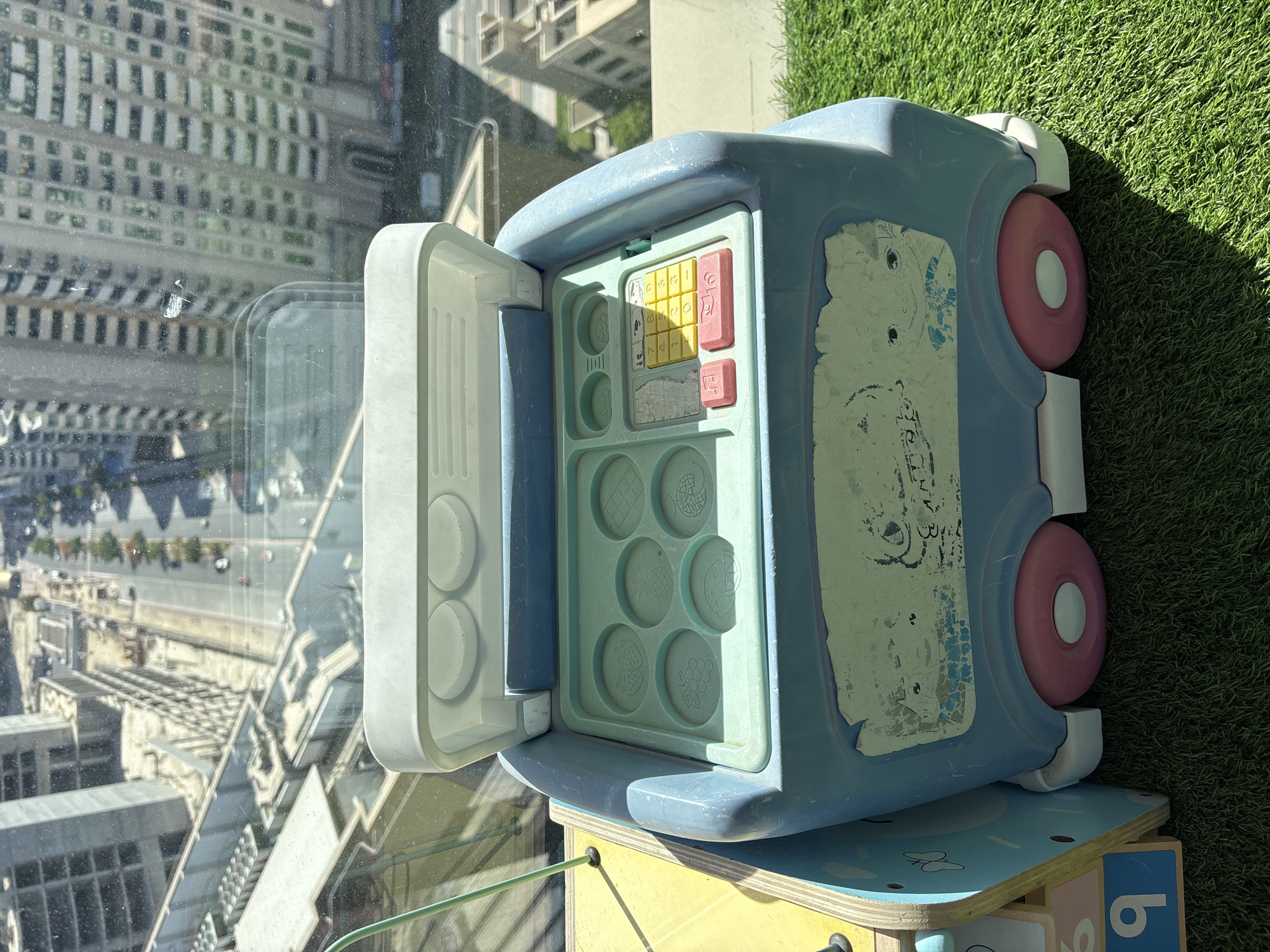A blue and white toy ice cream truck is positioned on a patch of artificial turf, in front of a large window that overlooks a city skyline. The window reveals a bustling urban environment, with skyscrapers and construction sites characteristic of Dubai, United Arab Emirates. The toy truck, which appears to be a cash register and ice cream set, has an open lid revealing compartments for ice cream shapes and a keypad with buttons. The exterior of the truck is worn, with faded decals and peeling paint. In the background, through the window, construction cranes and unfinished buildings suggest ongoing development. The scene is lit by sunlight, casting shadows on the turf.