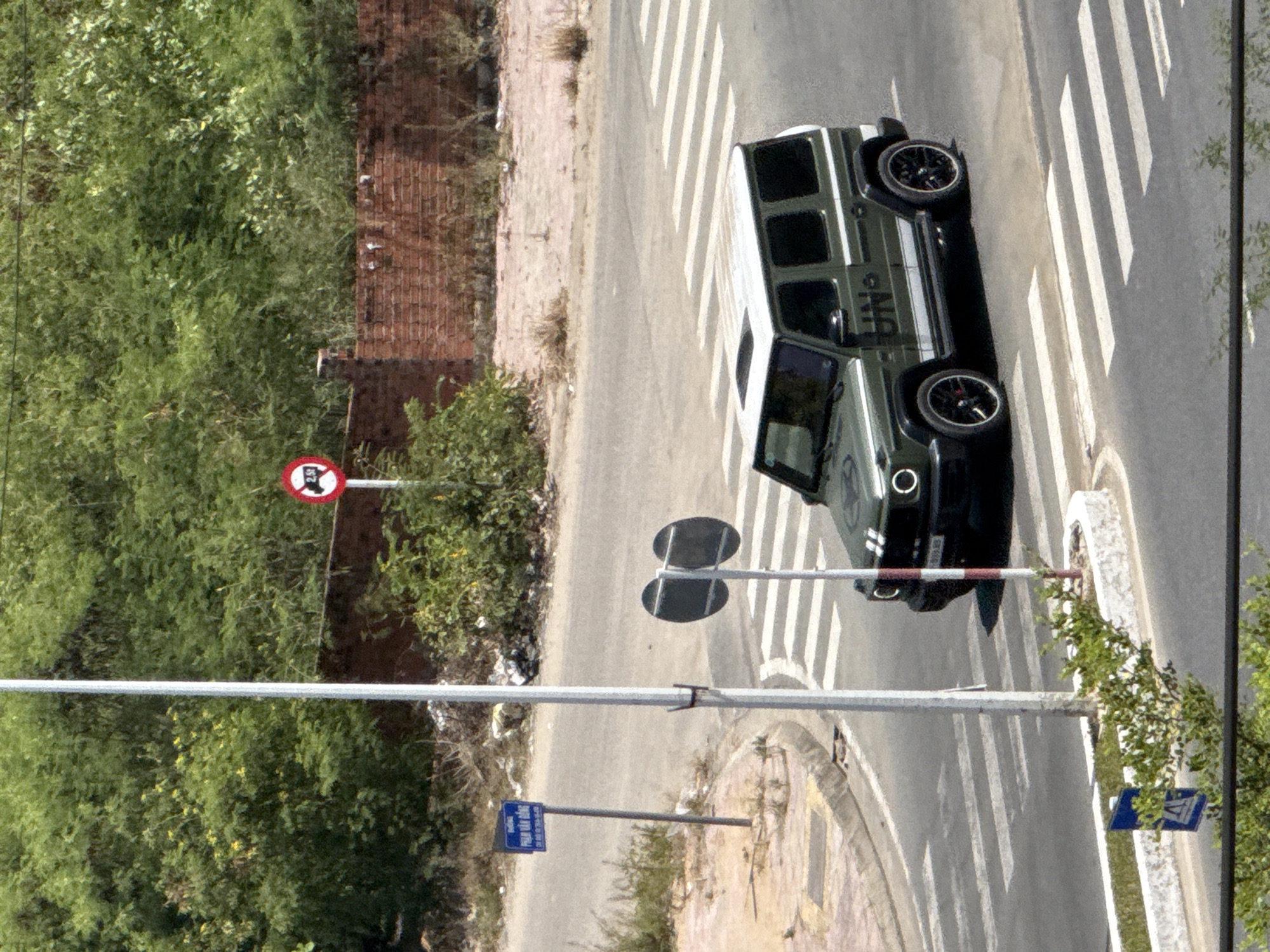 The image depicts an outdoor scene, likely in Nha Trang, Vietnam, given the context. The primary subject is a dark green Mercedes-Benz G-Class parked on a pedestrian crossing. The car has its doors open, suggesting it may have recently stopped or is about to move. The vehicle is adorned with white racing stripes and lettering that reads "AMG". Visible text on the car includes "AMG".
The setting is a paved road with clear white pedestrian markings. Adjacent to the road is a brick structure and lush green foliage, indicating a partly natural, partly urban environment. A red and white circular sign with a diagonal bar, likely a prohibition sign, is visible on the left side of the image, and its text is illegible. There are also traffic signals with dark circular plates. Another blue sign with Vietnamese text is present in the lower portion of the image.
The lighting suggests it is daytime, possibly sunny, given the shadows. The overall scene appears to be a still moment, with no people actively engaged in an event. The focus is on the vehicle and its immediate surroundings, highlighting the car's presence on the crosswalk.