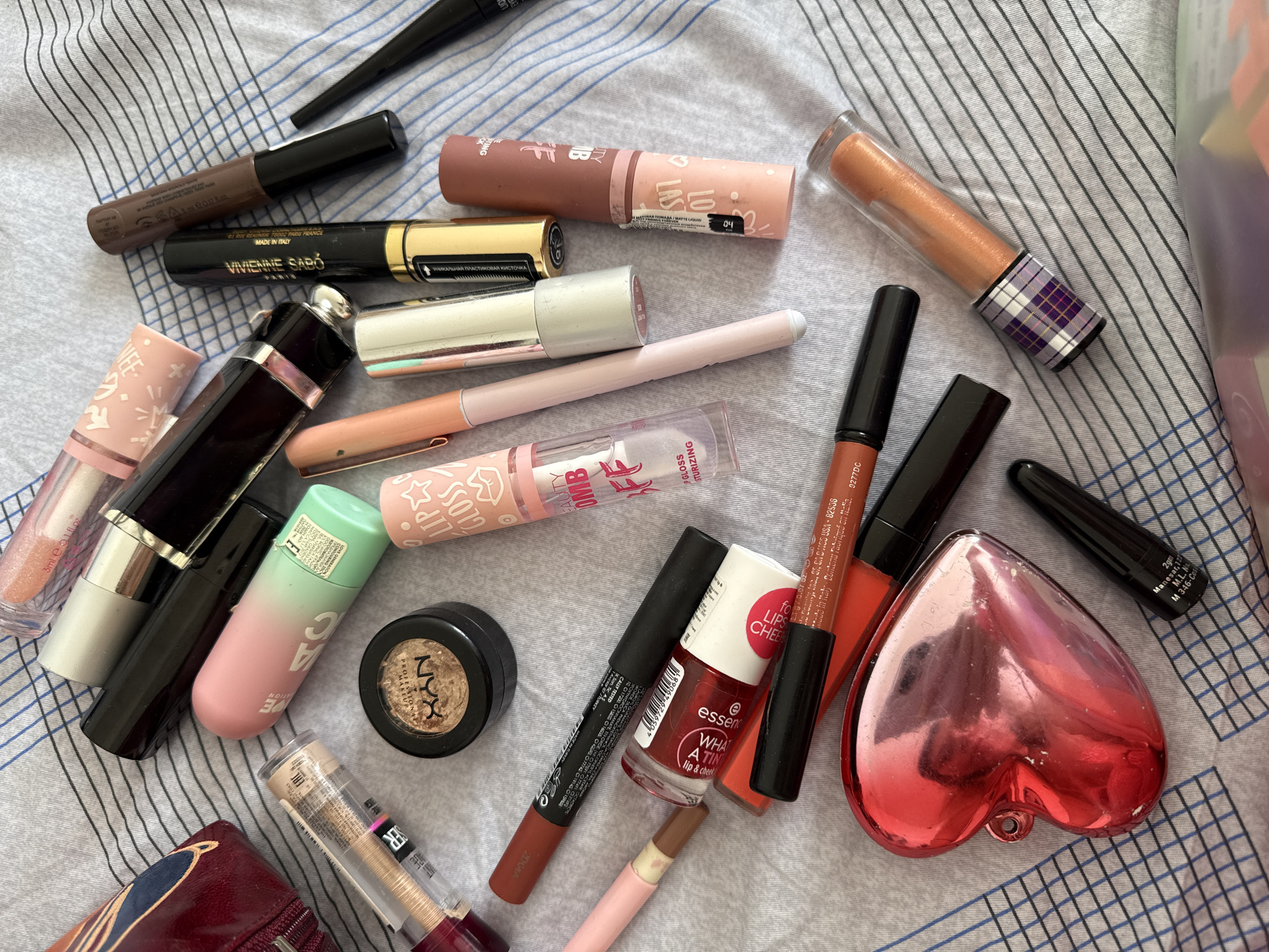 The image shows a collection of various makeup products scattered on a striped fabric surface. The products include lip glosses, lipsticks, eyeliners, eyeshadow, and nail polish from brands like NYX, Vivienne Sabó, Clinique, and Essence. Some products have visible labeling with locations, including Paris, France and Manesar, India. There is no indication that this image was taken in the city of Moscow, Russia.