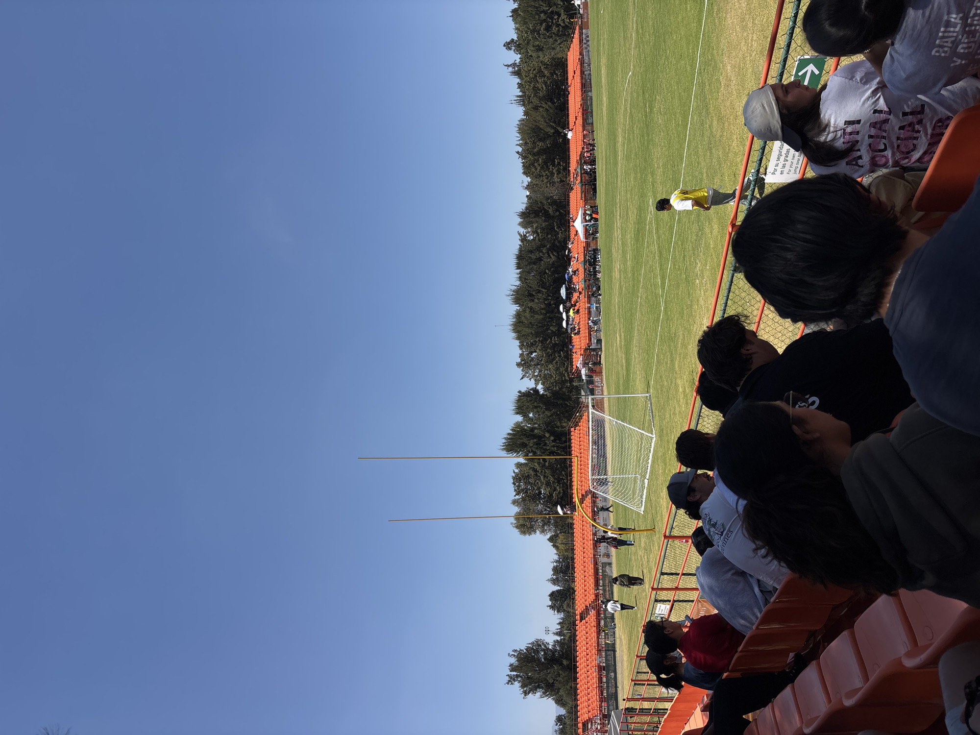 This image depicts an outdoor sports stadium in San Andrés Cholula, Mexico, under a clear, unobstructed blue sky. The foreground shows the backs and profiles of approximately 10-15 individuals, primarily young adults, seated in orange plastic stadium seats. One spectator wears a grey baseball cap and a white t-shirt with "ANTI SOCIAL SOCIAL CLUB" printed on the back. A green chain-link fence topped with an orange rail separates the spectators from the field. Attached to this fence is a white sign with a green arrow and Spanish text, partially obscured, which reads "Por su seguridad en las gradas / For your own safety on the stands."
The field itself is covered in well-maintained green grass with white markings, indicating a sports playing area. On the far left of the field, a white metal soccer goal with a net is positioned. To the right of the soccer goal, two tall, yellow goalposts, consistent with American football, stand vertically; one has a small white object attached near its base. Several individuals are present on the field: one male in a yellow shirt and grey pants stands near the right sideline, facing away from the camera, while approximately 5-7 other individuals walk near the soccer goal.
Beyond the field, a long section of orange plastic stadium grandstands stretches horizontally, appearing largely unoccupied in this section. Along the base of these far grandstands, several white canopies or tents are erected, under which small groups of people are visible. A dense line of dark green trees forms the perimeter background behind the grandstands and the event area. The scene is illuminated by direct sunlight, indicating daytime.