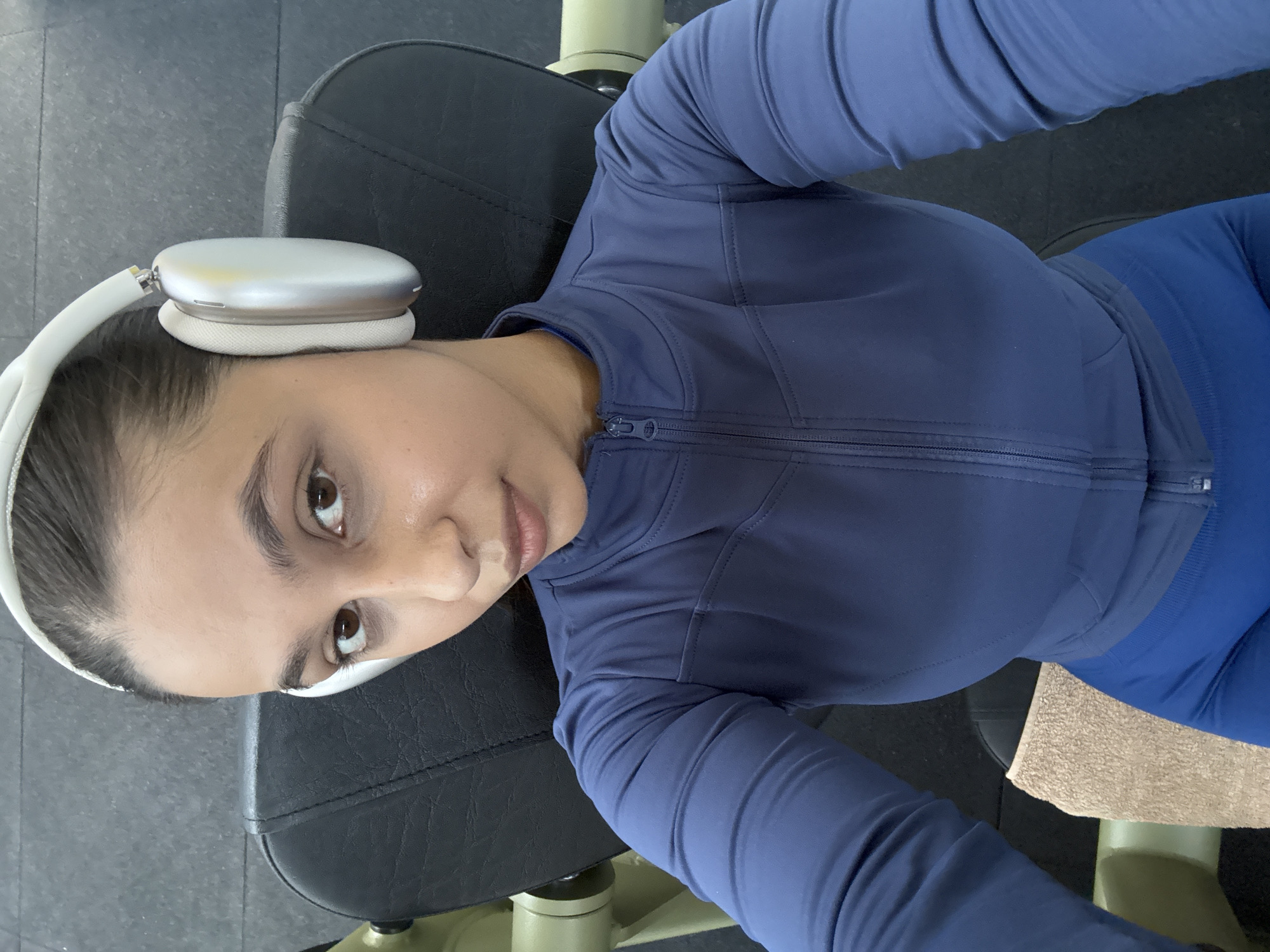 A woman lies on a black workout bench, wearing a blue athletic zip-up top and blue leggings. She has white headphones on. The scene appears to be inside a gym. There's a beige towel next to her. There is no indication of this picture's location in the city of Mönchengladbach, Germany.