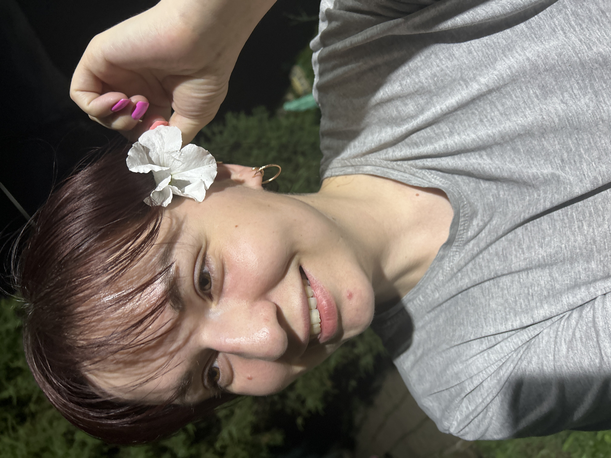A medium shot shows a young woman from the chest up, outdoors at night. She has short, dark reddish-brown hair and is wearing a grey t-shirt. She is smiling and looking to her left. A small white flower is tucked behind her right ear, and her right hand is reaching up to hold it. She has bright pink fingernail polish. A gold hoop earring is visible in her left ear. The background is dark, with some out-of-focus green foliage suggesting trees. The lighting appears to be artificial and casts a slight shadow on her face. The scene does not contain any specific identifying landmarks that would place it in the city of Fokino, Russia.