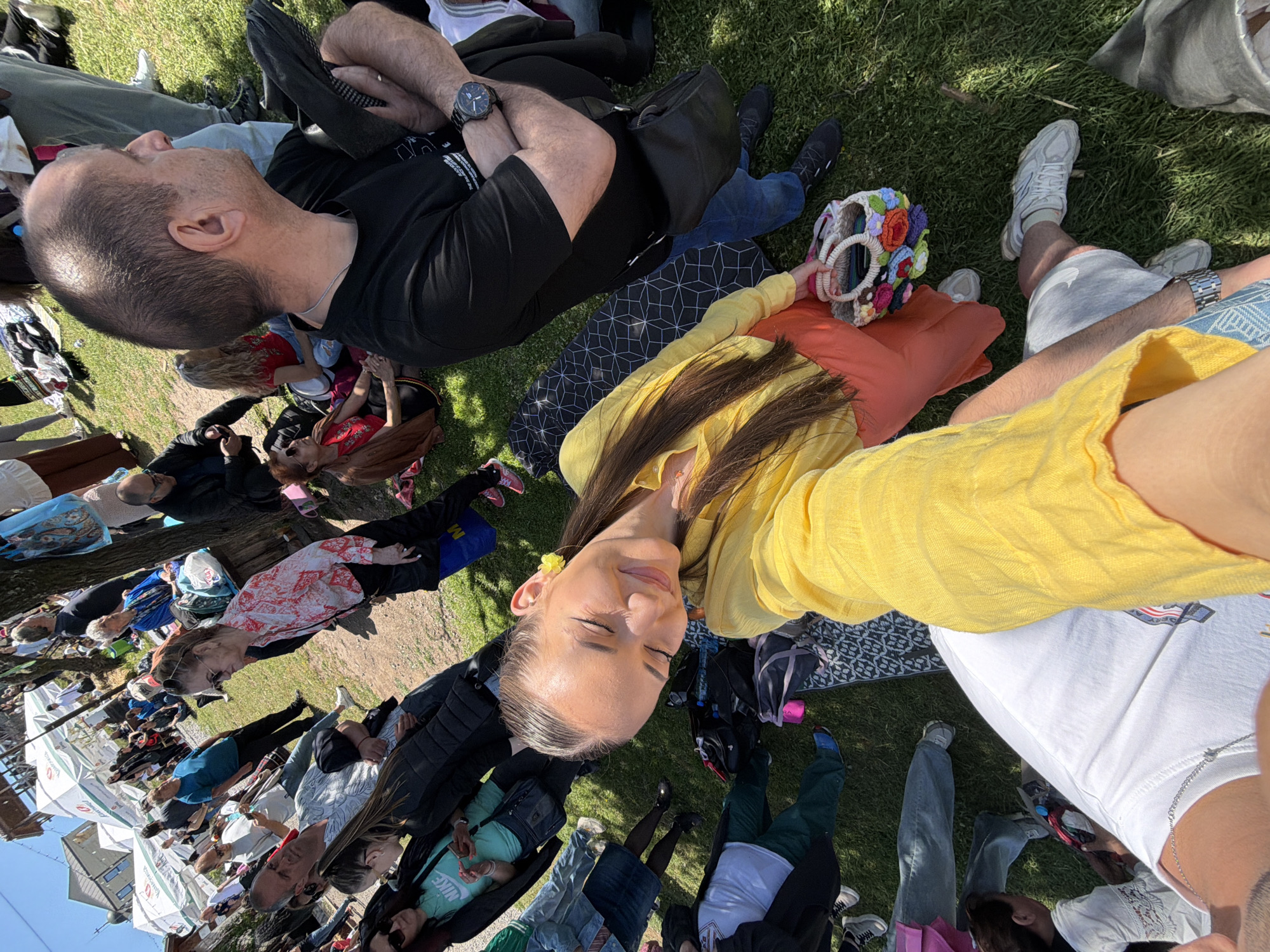 The image shows a large group of people sitting on a grassy area, presumably in a park. The perspective is from above, looking down at the scene. At the top of the frame is a man with a shaved head, wearing a black t-shirt. He is sitting with his arms crossed. Beside him is a colorful, decorative bag.
The foreground of the image features a woman wearing a yellow top. Other people are scattered around, many sitting on blankets or directly on the grass. There are also several bags and other personal items visible. The background includes trees and buildings.
The scene takes place in the city of Sofia, Bulgaria.