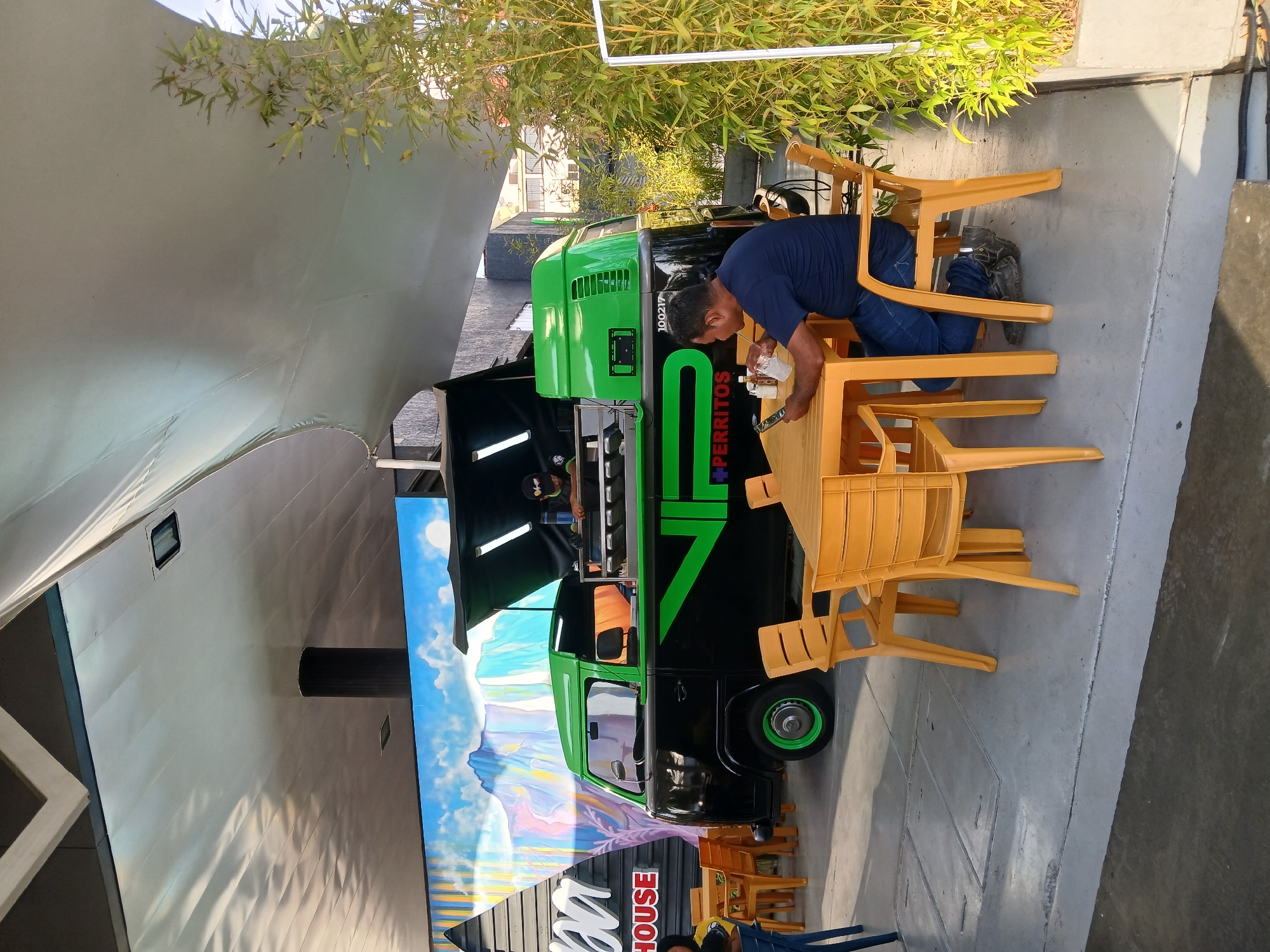 A man in a blue t-shirt and jeans stands leaning over a stack of orange plastic chairs next to a green and black food truck. The truck has "PERRITOS" in green letters on its side. The truck is parked in front of a building with a mural of mountains and clouds. A second person is visible in the truck. There are trees overhead. The location is in Ciudad Guayana, Venezuela.