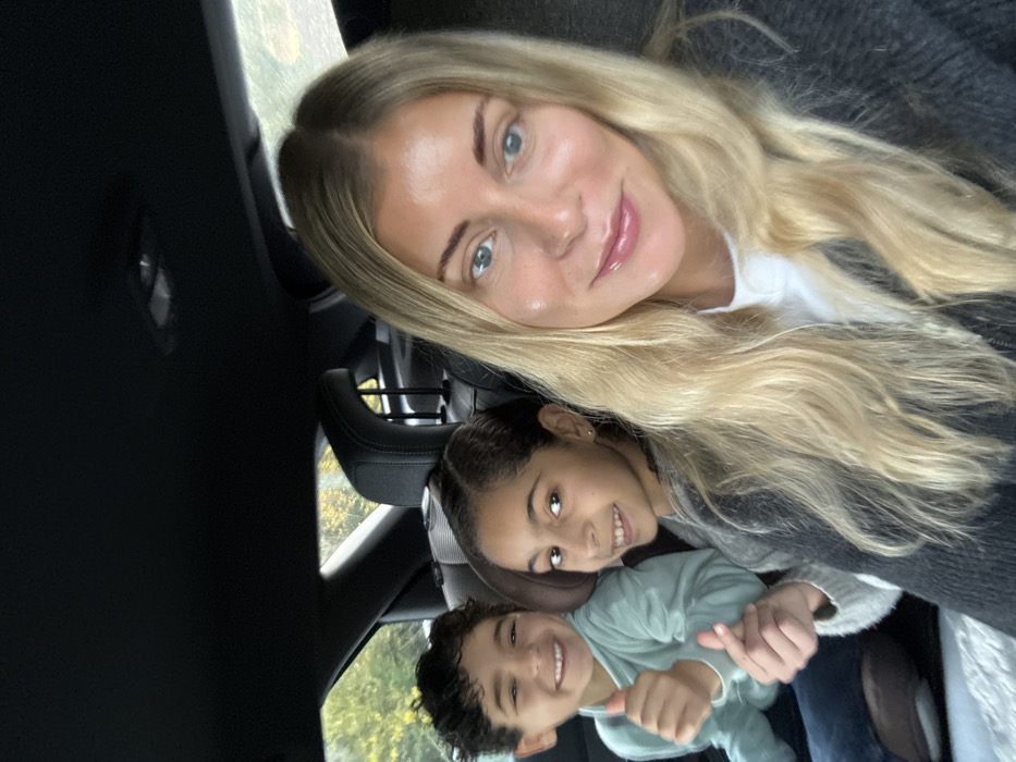 The image captures three individuals inside a vehicle, identified as being in Pourrières, France. In the foreground, an adult female with long blonde hair and blue eyes, wearing a white shirt under a dark gray cardigan, looks towards the camera with a slight smile, suggesting she is taking a selfie. In the back seat, two children are visible. On the left, a boy with dark curly hair, wearing a light blue-green long-sleeved top, smiles broadly and gives a thumbs-up gesture with his left hand. Next to him, a girl with dark hair, wearing a light gray sweater, smiles and forms a finger heart gesture with her right hand. Both children appear seated in the rear, looking at the camera. Through the car's rear window, blurred green and yellow foliage is visible, indicating an outdoor setting. The car interior features dark upholstery and headrests.