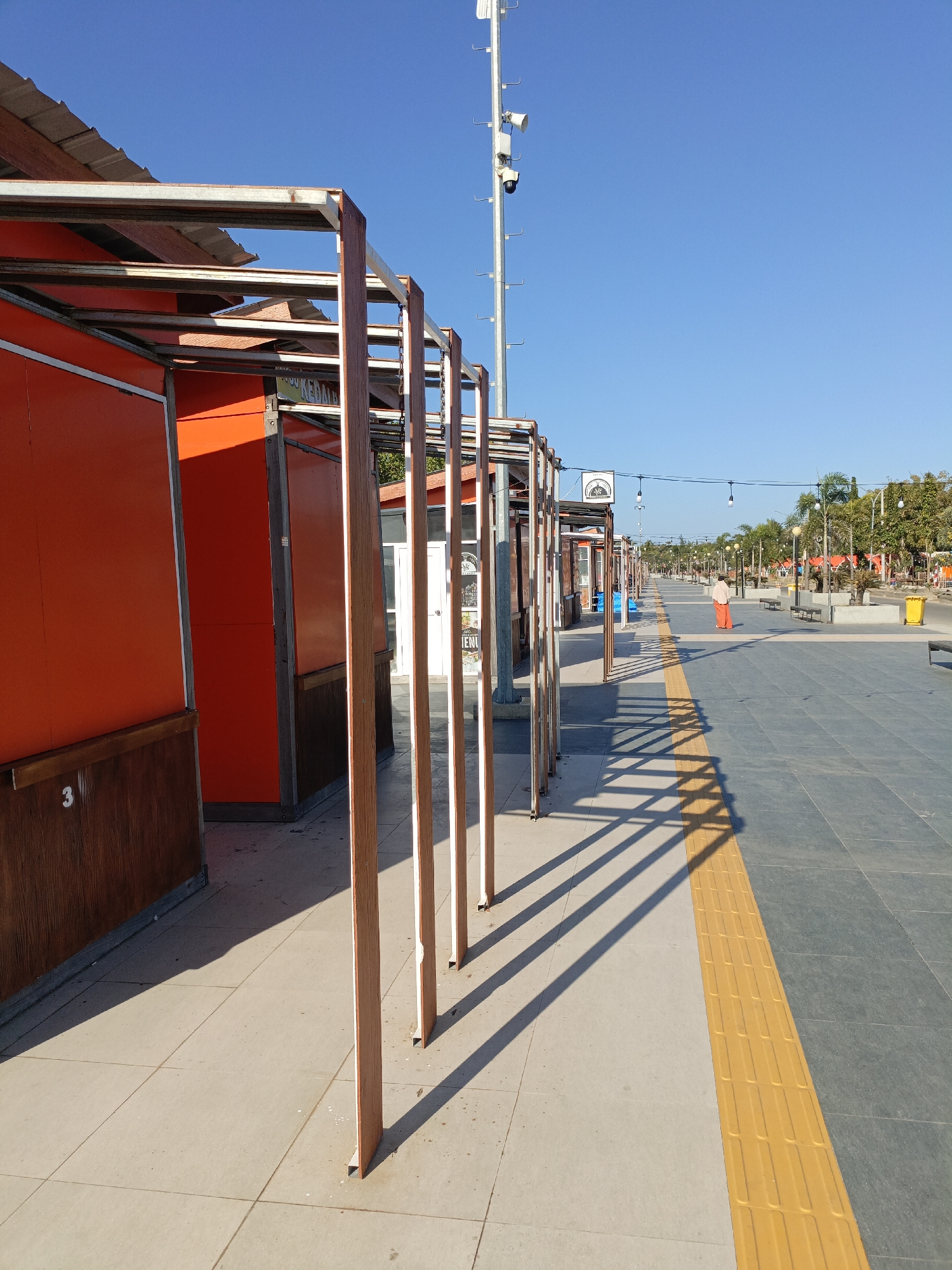 In the city of Ela-ela, Indonesia, a bright, sunny day illuminates a pedestrian walkway. The walkway is lined with orange kiosks on the left, featuring wooden lower panels. A metal frame structure extends from the kiosks, casting long, geometric shadows on the pale tiled ground. Further down the walkway, a yellow tactile paving strip runs alongside the dark grey pavement. A lone individual, wearing an orange garment, walks away from the camera towards the distance. The background shows a tree-lined avenue with more kiosks, lampposts, and a tall mast with cameras and speakers, indicating a public space designed for foot traffic and surveillance. The sky is a clear, vibrant blue.