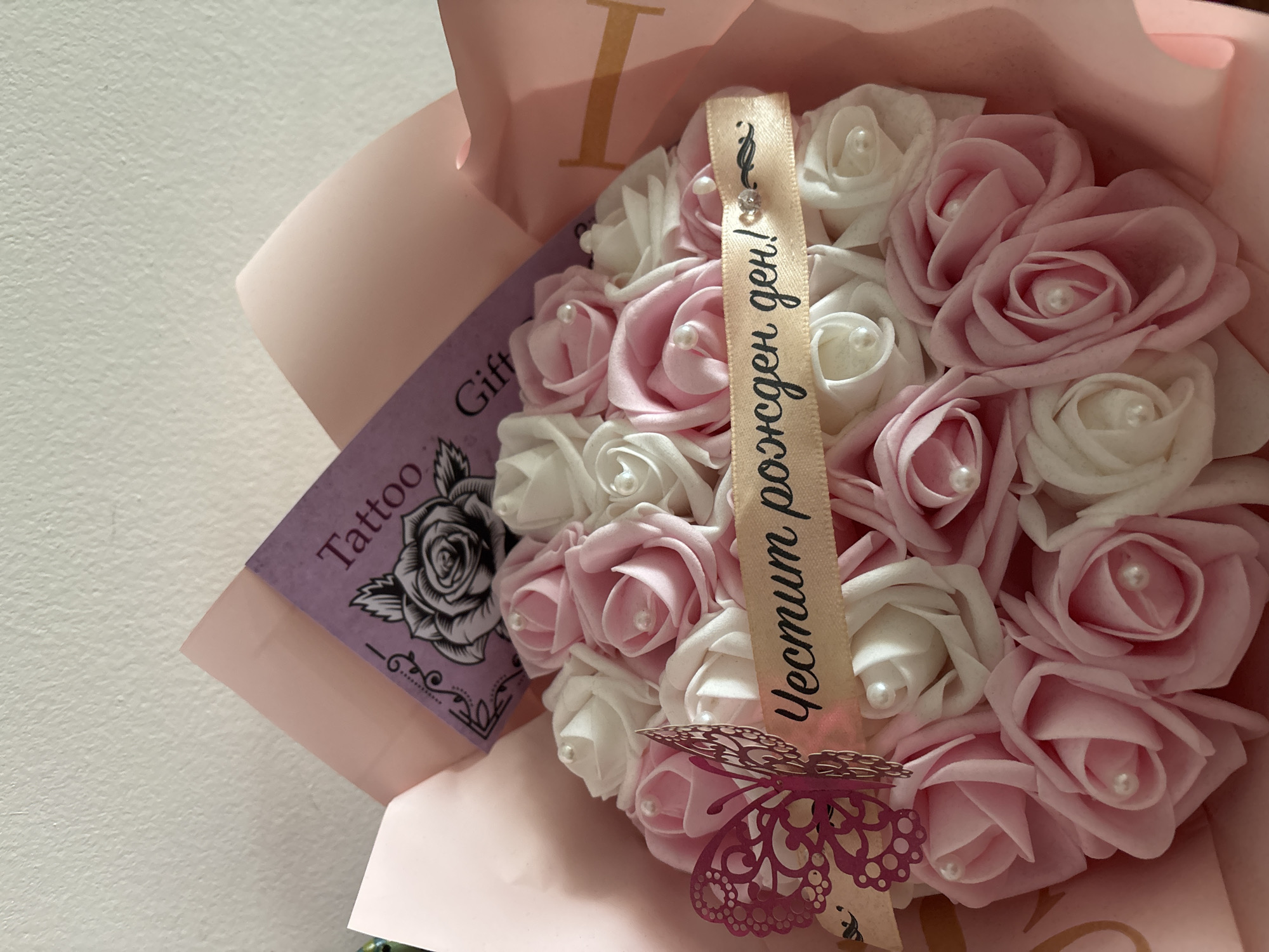 This image captures a bouquet of artificial roses in a soft pink and white color scheme, each rose featuring a small pearl in its center. The bouquet is wrapped in light pink paper or fabric and adorned with a beige ribbon. The ribbon displays text in Cyrillic, "Честит рожден ден!", which translates from Bulgarian to "Happy Birthday!". A decorative, cut-out style pink butterfly is attached to the ribbon near the bottom of the bouquet.
To the left, a purple gift tag is partially visible, showing the English words "Tattoo Gift" alongside a detailed illustration of a rose. The background appears to be a light-colored, textured wall, suggesting an indoor setting. The overall presentation indicates a gift for a special occasion, likely a birthday celebration, taking place in Troyan, Bulgaria. The lighting is bright, highlighting the delicate details of the artificial flowers and decorations.