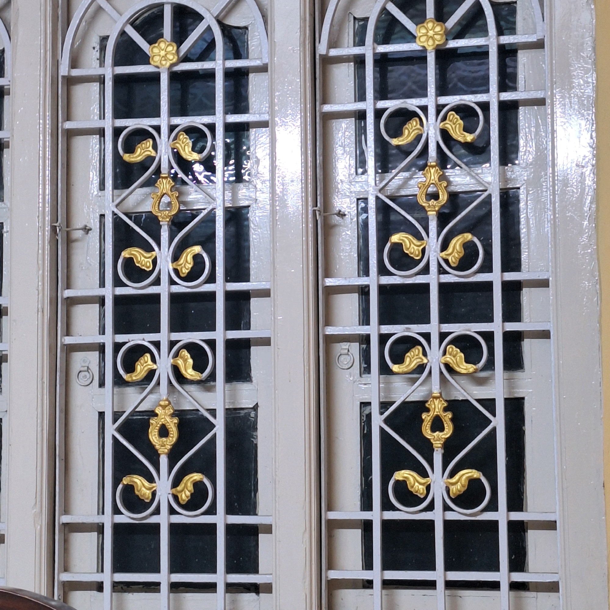 The image shows an outdoor view of a closed wooden door with ornate metal grilles. The door is painted a light color, possibly cream or white, with visible brushstrokes and some wear. Each side of the door features a decorative grille composed of vertical and horizontal bars, with arched tops. These grilles are adorned with golden floral and leaf-like embellishments. The right side of the door has a small metal latch. The background visible through the grilles appears to be dark, suggesting the interior is not brightly lit. The overall impression is of an older architectural style. The location is specified as Gudrukoppa, India.
