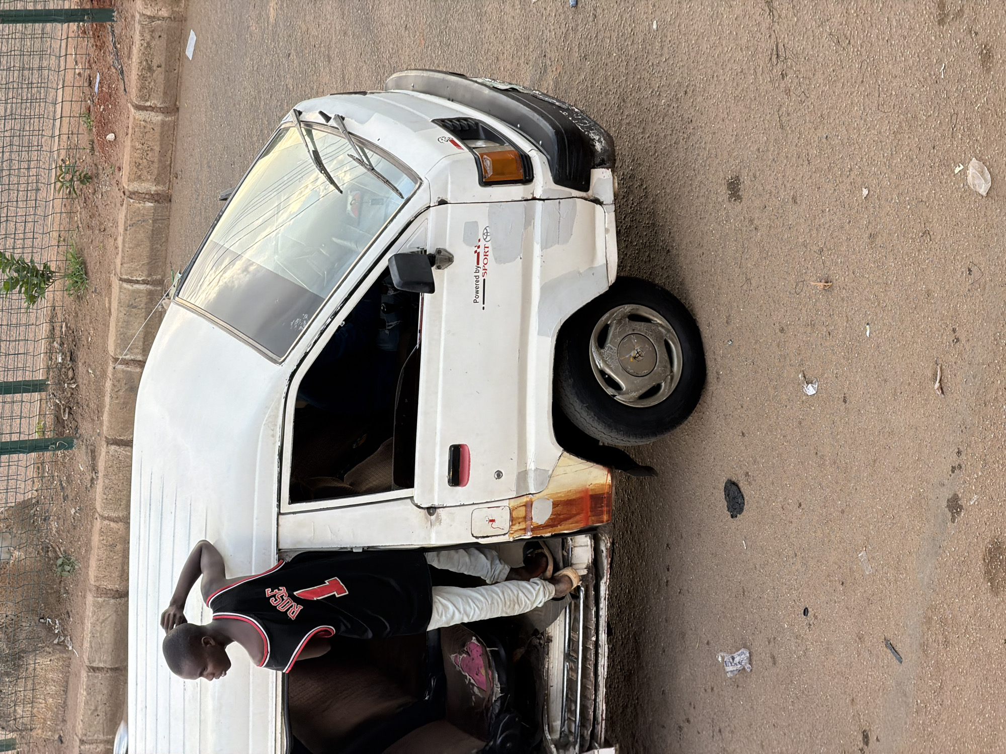 In Bukuru, Nigeria, a young Black male, appearing to be in his late teens, is partially inside a white van, leaning out of the open driver's side door. He is wearing a black basketball jersey with red trim, emblazoned with the number '1' and the name 'ROSE' across the back. His left arm is bent and resting on the roof of the van, while his right hand is near his head. He is wearing light-colored pants and brown sandals. The van is weathered, with visible rust on the lower portion of the door and evidence of repair on the front fender. The interior of the van appears worn, with visible seat upholstery. The van is parked on a paved road with a curb and sidewalk visible in the background. The area shows signs of being a busy street, with debris scattered on the road surface. A green metal fence with vegetation behind it lines the sidewalk.