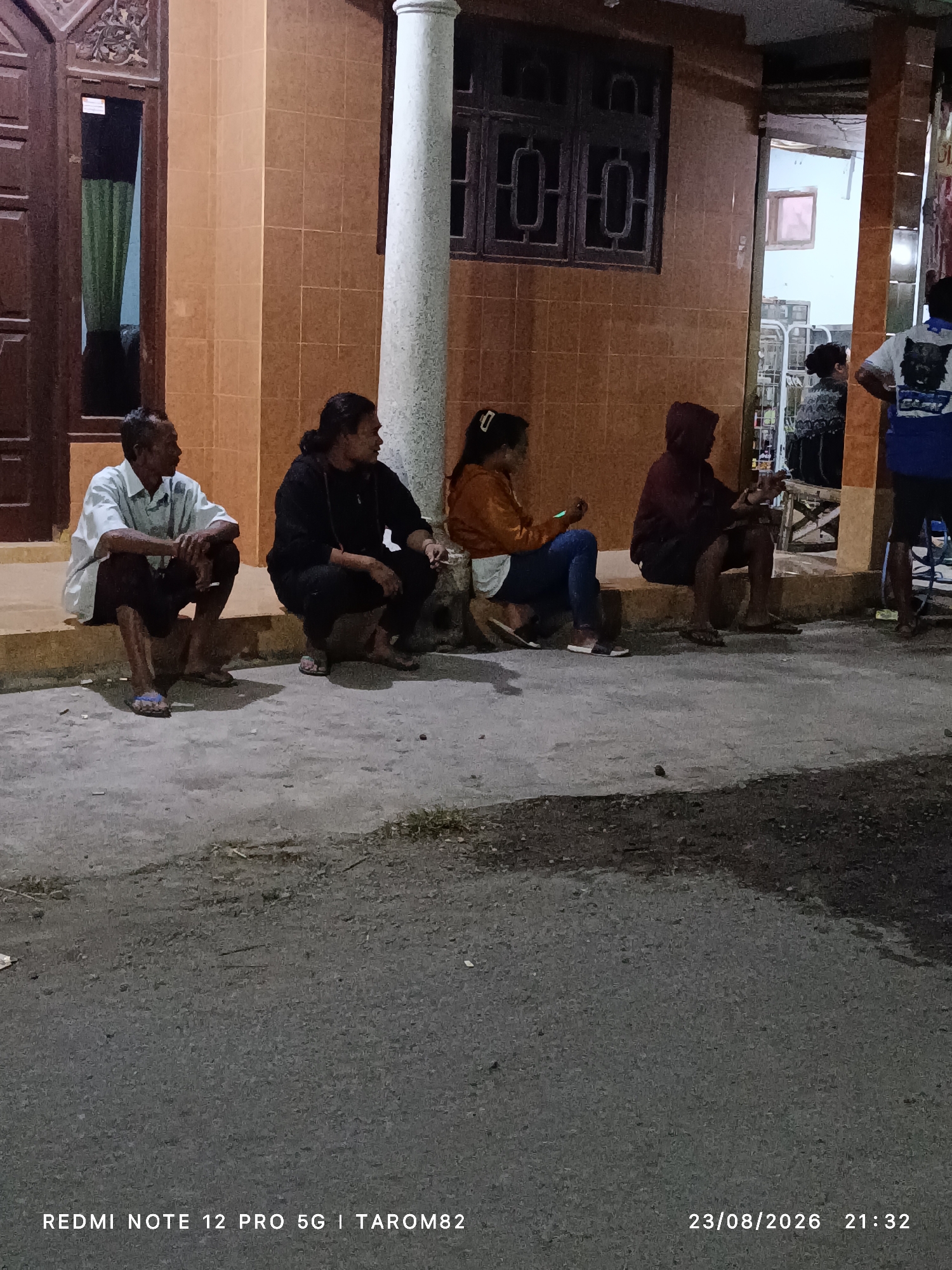 The image shows a group of five individuals sitting on the steps and sidewalk in front of a building in Rejoagung, Indonesia, at night. The building has orange tiled walls and a dark wooden door. On the left, a man in a white shirt and dark pants sits on the step, looking to his left. Next to him, a person in a black hoodie and dark pants is also seated, looking to their left. Further to the right, a person in an orange jacket and blue jeans sits on the step, looking at a mobile phone. Another person in a dark hooded jacket and dark shorts sits on the step, facing right. On the far right, a person in a blue and white t-shirt and dark shorts stands, facing left. The ground in front of the building is a paved surface. The overall lighting suggests it is evening or night. A watermark at the bottom indicates the date "23/08/2026" and time "21:32".