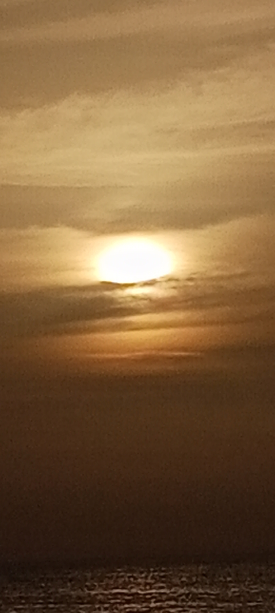 The image depicts a sunset over the ocean, likely in Juan Griego, Venezuela. The sun, a bright, luminous disc, is partially obscured by thin, hazy clouds, casting a warm, golden glow across the sky. The clouds themselves are layered, with some appearing lighter and more diffused, while others form darker, horizontal bands. Below the sky, the ocean is visible as a dark, shimmering expanse, with subtle ripples reflecting the dim light. The overall impression is one of a serene and tranquil twilight scene, characteristic of coastal sunsets.
