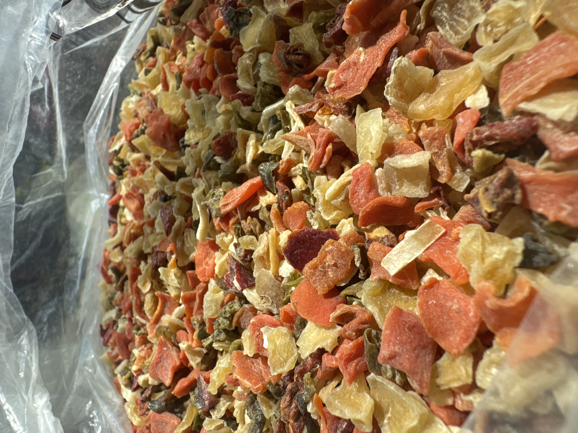 A close-up, low-angle, eye-level shot captures a large quantity of dried fruit pieces, likely from within a clear plastic bag. The pieces are small and irregularly shaped, with a mix of vibrant colors including orange, yellow, and deep red, interspersed with some greenish-brown elements. The texture appears somewhat rough and dehydrated. The top of the plastic bag is visible, creating a wrinkled, translucent barrier that casts shadows and diffuses light across the contents. The background is largely obscured by the plastic and the sheer volume of the dried fruit, with indistinct gray and bright areas suggesting the presence of light and shadow. The overall impression is a detailed view of a bulk food item. There are no people or specific environmental details within a city.