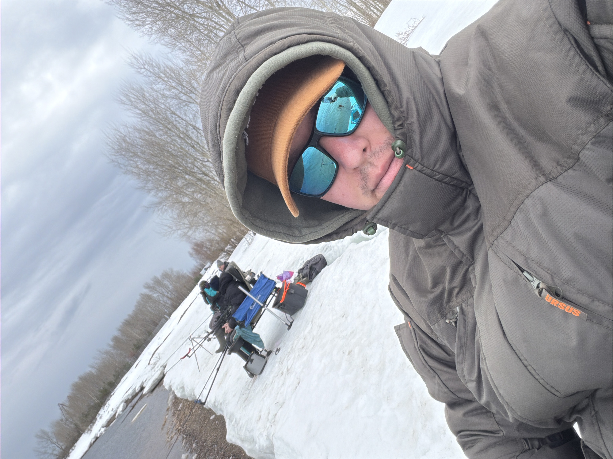 The image depicts a selfie taken by an individual wearing a brown hooded jacket, a tan baseball cap, and blue reflective sunglasses. The jacket has the word "URSUS" visible on a pocket. In the background, a snowy landscape is visible, with a group of people fishing. Fishing rods are set up along the edge of a body of water. The location is in Chaikovsky, Russia.