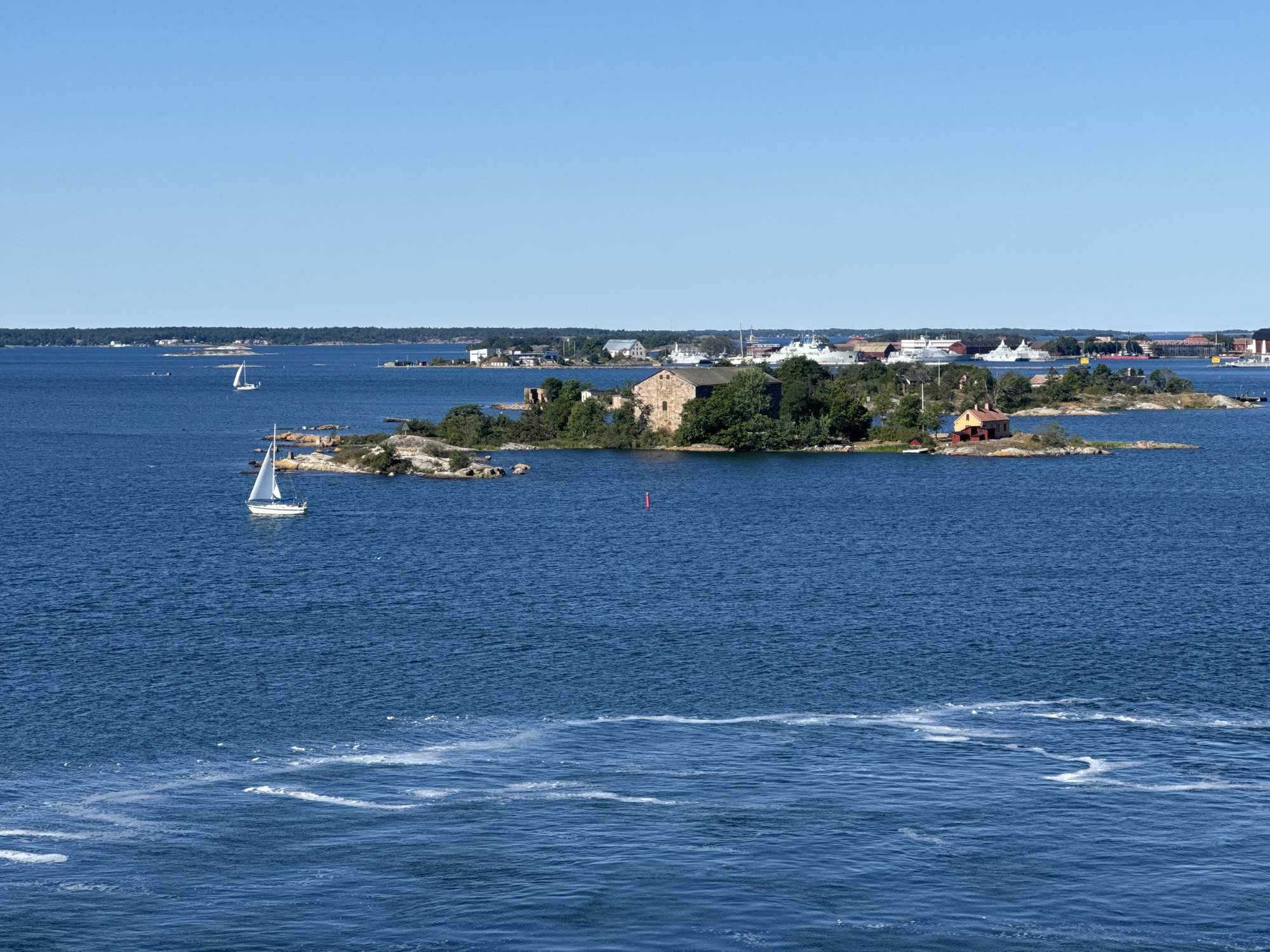 The image depicts a bright, sunny day with a clear blue sky over a body of water. In the foreground, the deep blue water exhibits some white foam trails, suggesting movement.
In the middle ground, a small, rocky island is visible, densely covered with green trees. On this island, there is a rustic stone building, appearing to be a ruin, and a small, orange-roofed building with red walls. Further to the right, another small island features a single red-roofed house.
Beyond the islands, the water extends to a distant coastline. On this coastline, various buildings and structures are visible, including what appear to be docks and several large ships, some of which have white hulls and superstructure. The scene suggests a port or naval base area.
Two sailboats are present in the water. One is closer to the foreground, a white sailboat with its sail up, moving from left to right. Another smaller white sailboat is visible further in the distance, near the islands. A red buoy is also present in the water.
The overall impression is of a picturesque coastal landscape with a mix of natural beauty and human activity, likely related to maritime operations. The location is identified as the southern archipelago of Karlskrona, Sweden, a UNESCO World Heritage site known for its naval history and picturesque islands.