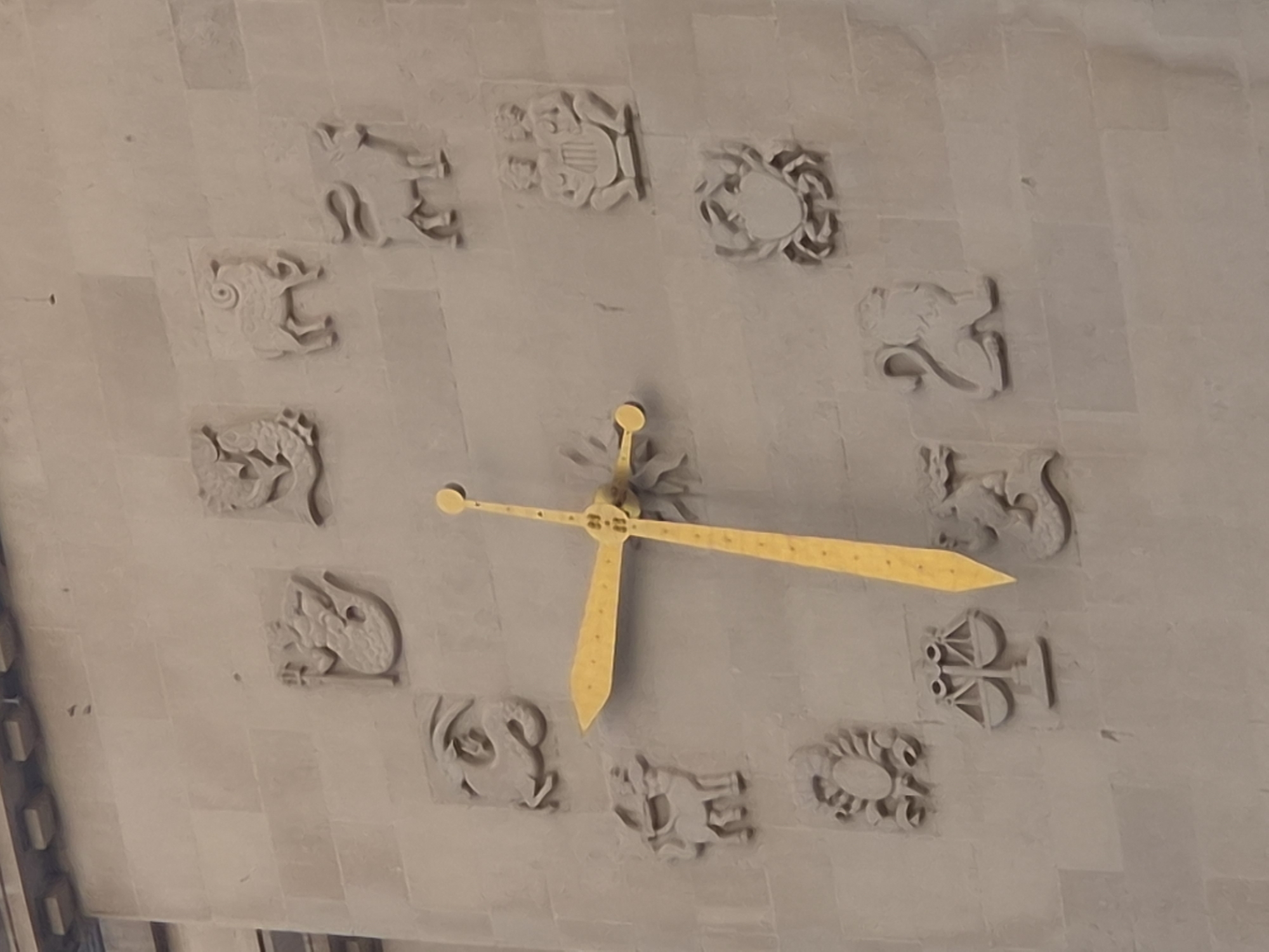 The image shows a clock on the facade of a building, likely a public institution, in Ostend, Belgium. The clock face is adorned with twelve relief carvings of astrological symbols, representing the signs of the zodiac. The clock hands are gold and appear to be positioned at approximately 2:10. The astrological symbols are arranged in a circular pattern around the clock's central mechanism. The building's facade is made of light-colored stone blocks. The image is taken from a slightly upward angle, suggesting the clock is mounted at a considerable height. The astrological symbols are carved in a stylized manner, with clear representations of each sign. The overall impression is one of historical and artistic decoration on an architectural structure.