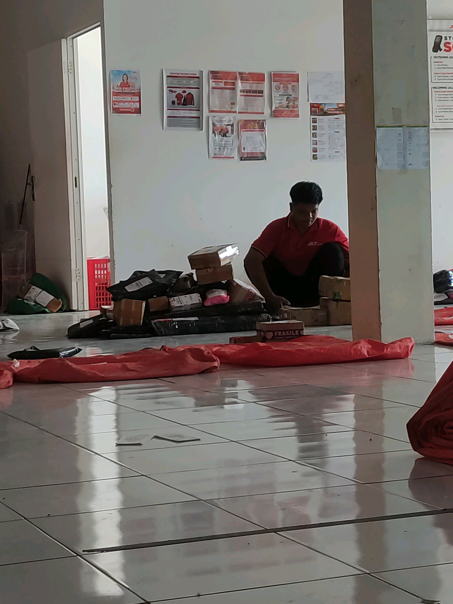 In a room with tiled floors, a man wearing a red J&T Express uniform crouches amidst a large pile of packages. The packages, of various sizes and shapes, are stacked haphazardly on the floor and include cardboard boxes and some wrapped in black plastic. Some packages are marked with shipping labels. The man appears to be sorting or organizing the packages. In the background, there is a doorway leading to another area, and posters are affixed to the wall. The lighting suggests an indoor setting, possibly a logistics or delivery hub in Rejosari, Indonesia.