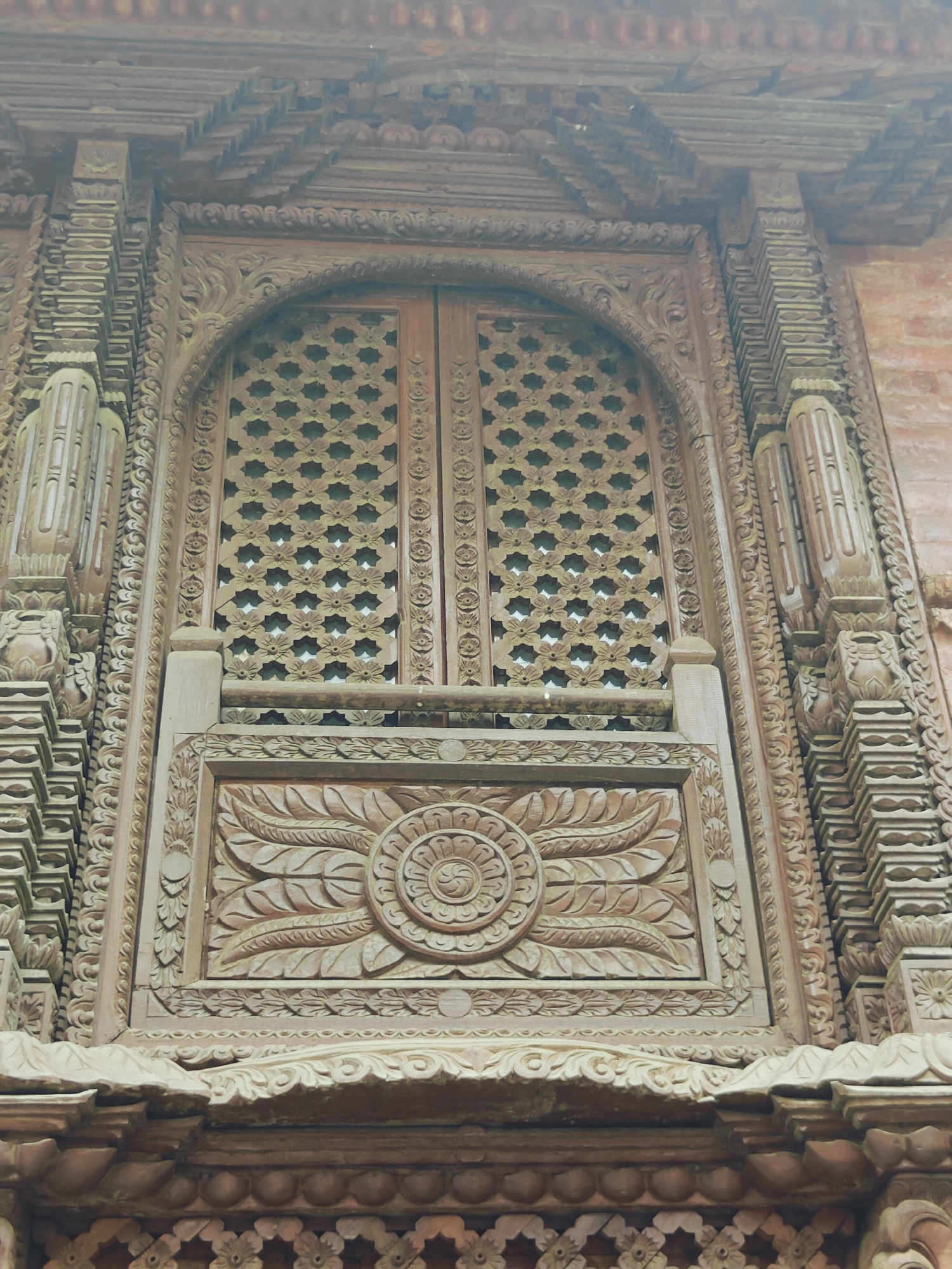 The image depicts an ornate wooden window with intricate carvings on the facade of a building. The window is set within a larger architectural structure featuring elaborate wooden detailing. Above the window, a series of layered wooden eaves are visible, and the surrounding walls appear to be made of brick. The central element is the large wooden window with two doors, each adorned with a repetitive pattern of star-like floral motifs. Below the window is a prominent wooden panel featuring a central circular motif surrounded by radiating leaf-like carvings. The entire structure showcases fine craftsmanship in woodworking. The building is located in the city of Kirtipur, Nepal. There are no people or animals visible in the image.
