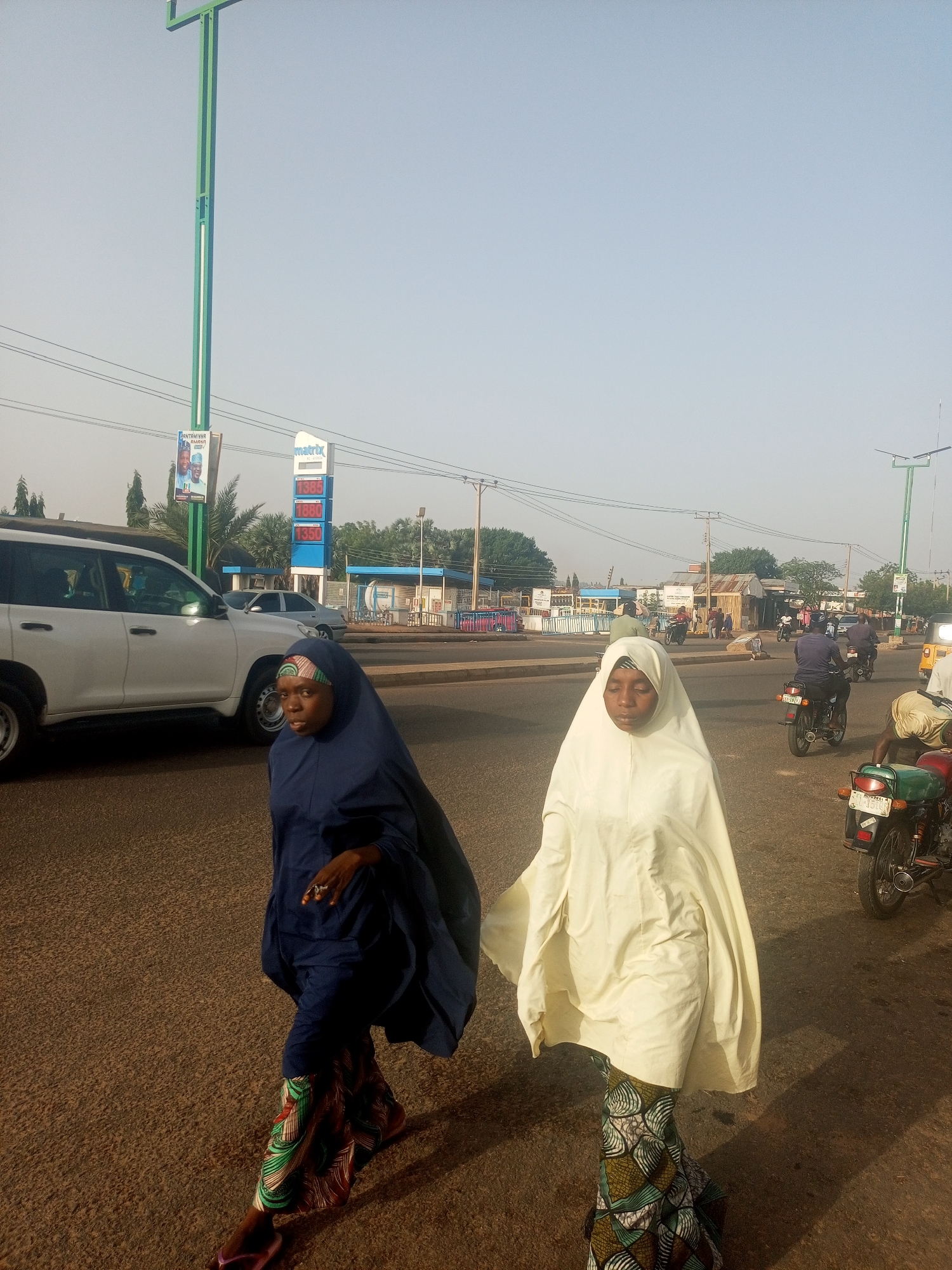Two women wearing hijabs and long patterned skirts are walking along a street in Garko, Nigeria. The woman on the left is wearing a navy blue hijab, while the one on the right is wearing a cream-colored hijab. There is a white SUV parked on the left side of the street. In the background, there is a gas station with a Matrix sign displaying gas prices. A few motorcycles and other vehicles are visible on the street and near the gas station. The sky is clear and light.
