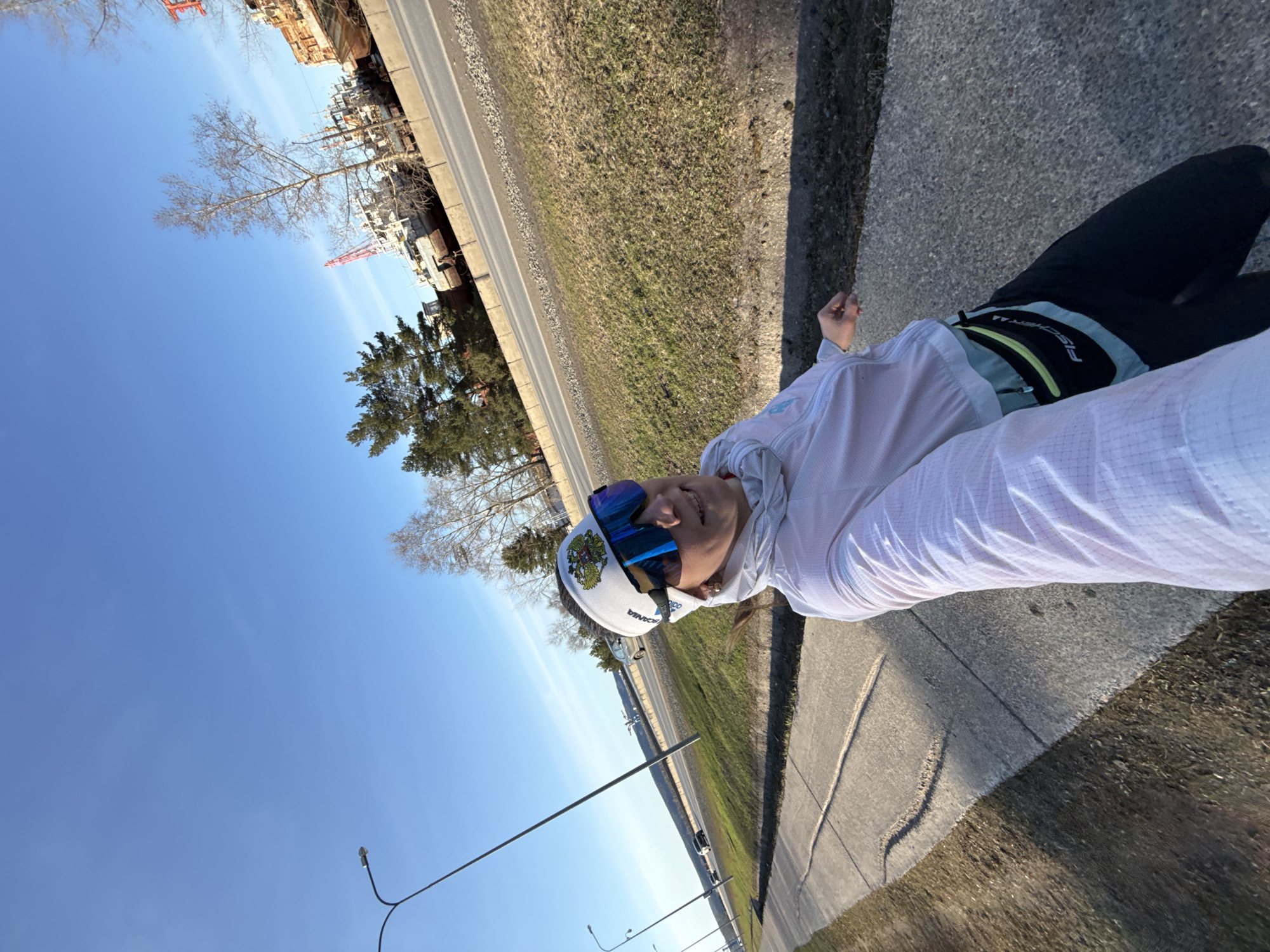 A person wearing a white shirt, black pants, a white Adidas hat with a Russian coat of arms, and blue-tinted sports glasses is standing on a sidewalk. They are holding a camera or phone in front of them, likely taking a selfie. The background includes a road with cars, a grassy area, trees, and buildings in the distance. Streetlights are visible along the road.
Based on the clothing and hat, the person may be Russian. Considering the mention of the city of Chaikovsky in Russia, it is reasonable to infer the photo was taken in or near that city.
