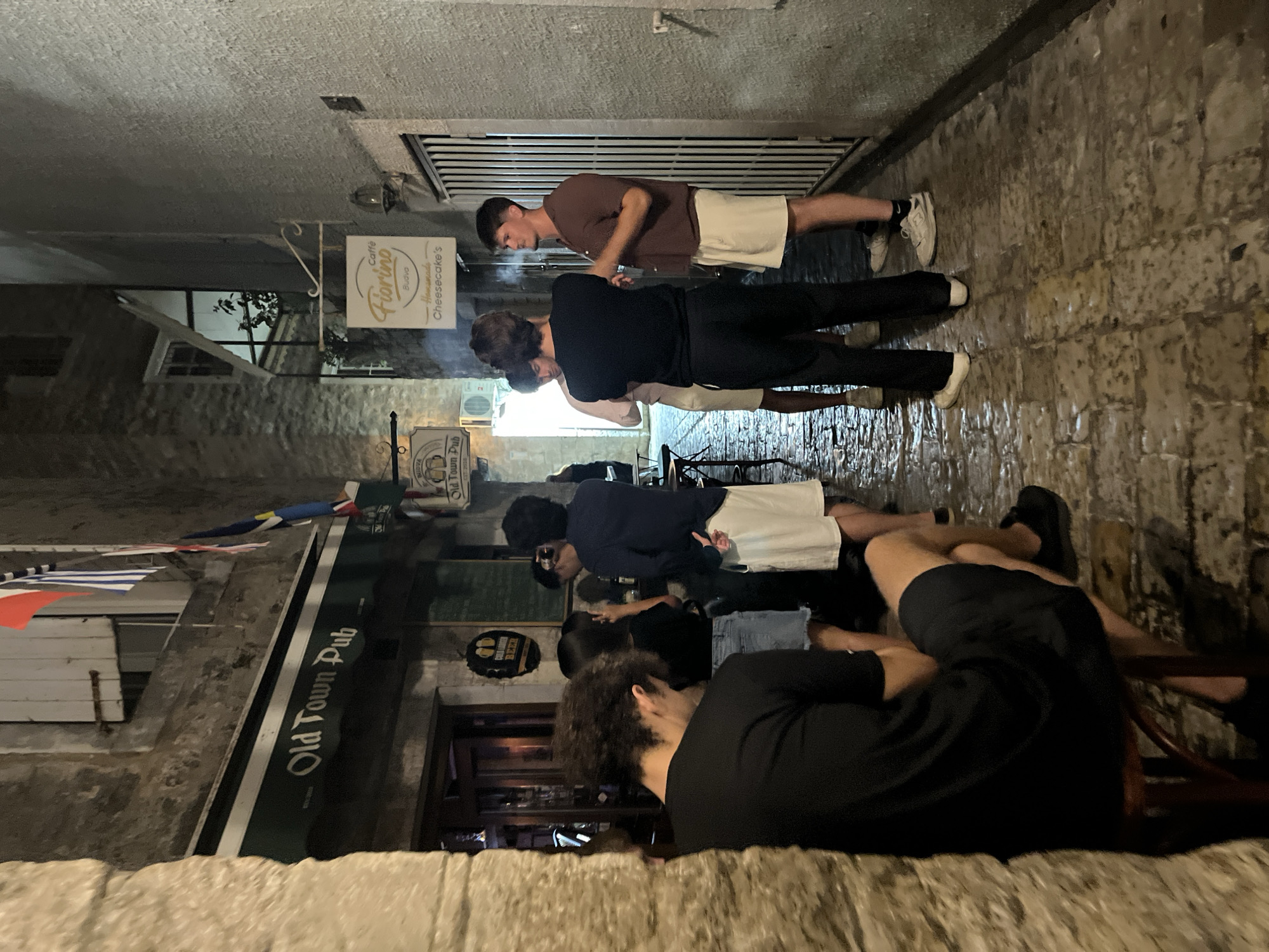 In Budva, Montenegro, a narrow, cobblestone alleyway is illuminated by artificial light. On the left, the facade of "Old Town Pub" is visible, featuring a green awning with white lettering. A few people are standing outside the pub, and a person with curly hair is seated on a stool in the foreground, facing away from the camera. Further down the alley, on the right wall, a sign for "Caffè Fiorino Budva - Homemade Cheesecake's" hangs. A group of four young men are standing and conversing in the middle of the alley, near the entrance to a building with a white metal gate. The alley floor is wet and reflects the light, suggesting recent rain. Flags are visible hanging from the buildings, including the Montenegrin flag.