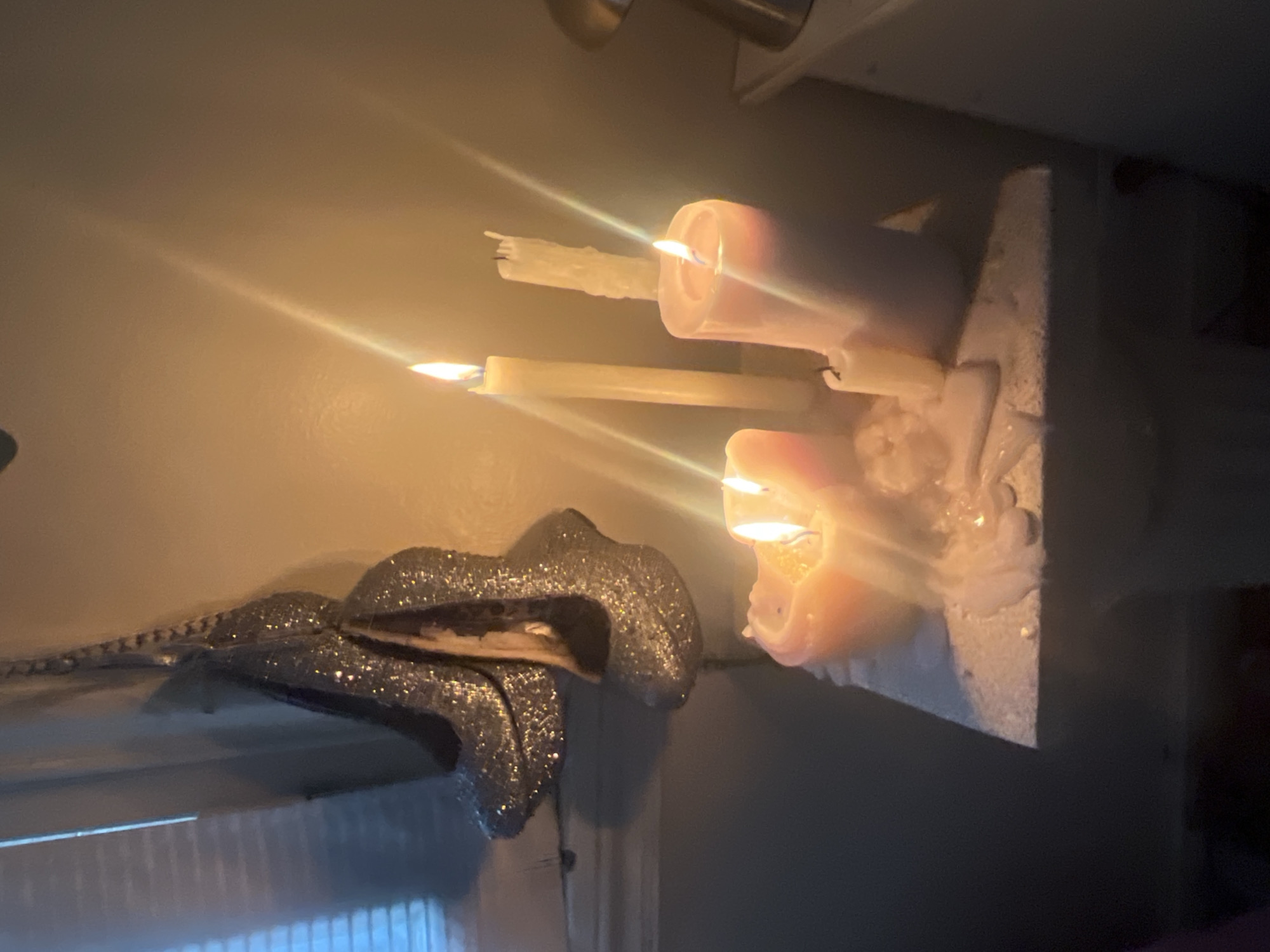 The image shows a still life composed of several objects. A collection of lit candles of varying sizes and shapes are mounted on what appears to be a stone or concrete base, with wax drippings covering the structure. Adjacent to the candles is a decorative object that resembles a pair of lips with glittery, reflective material covering its surface. The background is a lightly colored wall. The lighting suggests an indoor setting with the candlelight being the primary source. The location is unknown; there is no evidence in the image to suggest that the photo was taken in Buenos Aires, Argentina.