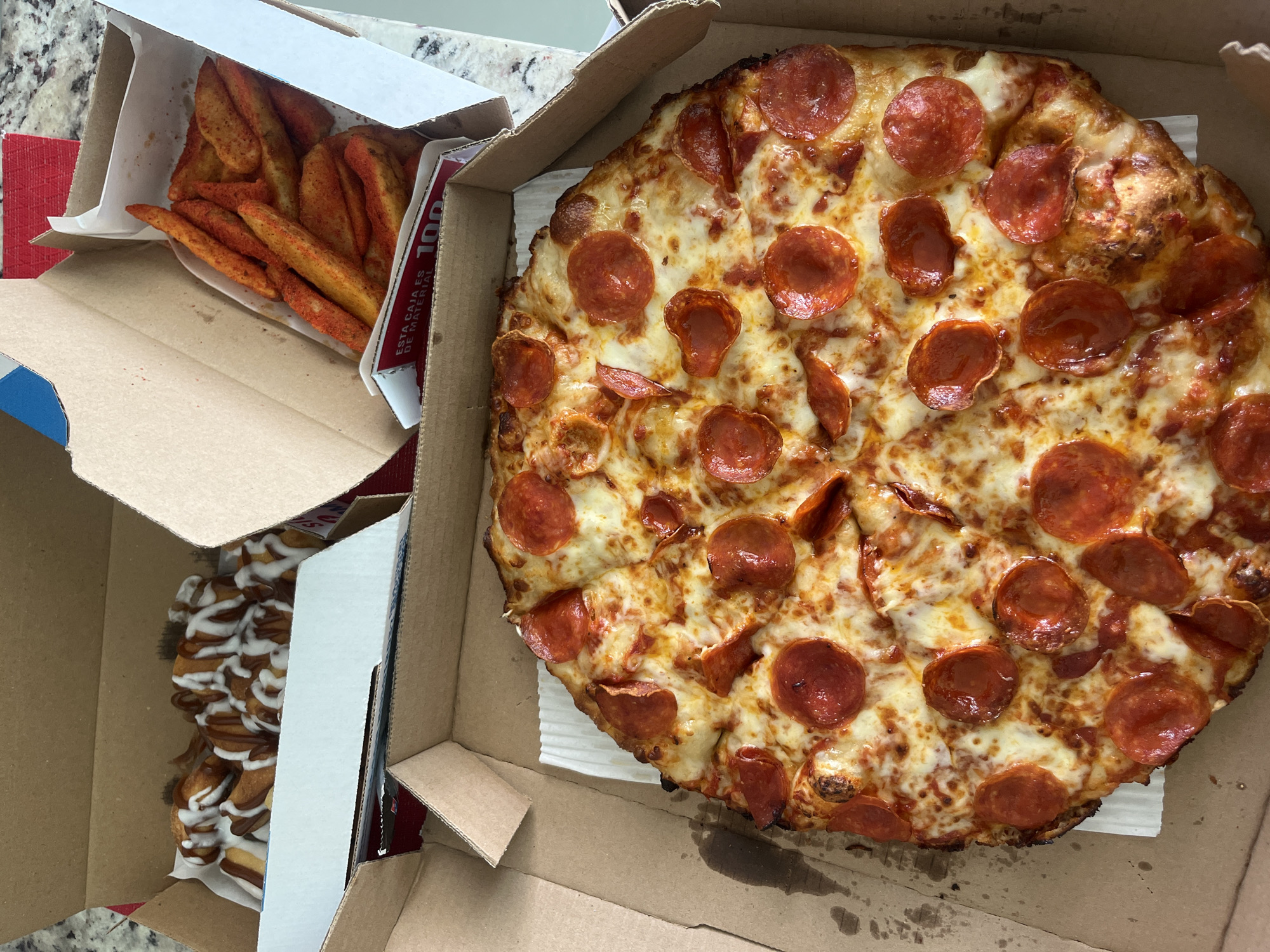 The image shows a partially opened pizza box containing a pepperoni pizza, alongside two other partially opened boxes. One contains potato wedges, and the other appears to contain dessert items with a drizzle of icing. The environment suggests an indoor setting, possibly a residence, with a countertop surface visible. There is no information within the image to suggest it was taken in the Barrio de Nuevo León, Mexico, nor is there information about people, actions, or interactions.