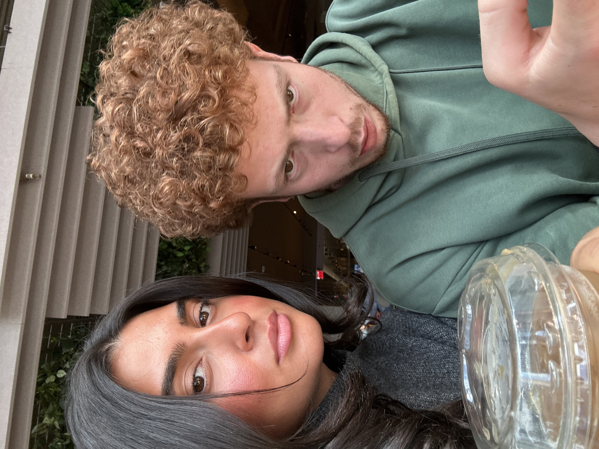 The image shows two people posing for a selfie. A man with curly auburn hair, wearing a green hoodie, is positioned at the top of the frame. A woman with long, dark hair is positioned at the bottom of the frame. A plastic cup is visible. The location appears to be in an indoor setting, potentially a cafe, in New York City, United States, as evidenced by the vertical building façade and the type of light fixtures visible through the window.