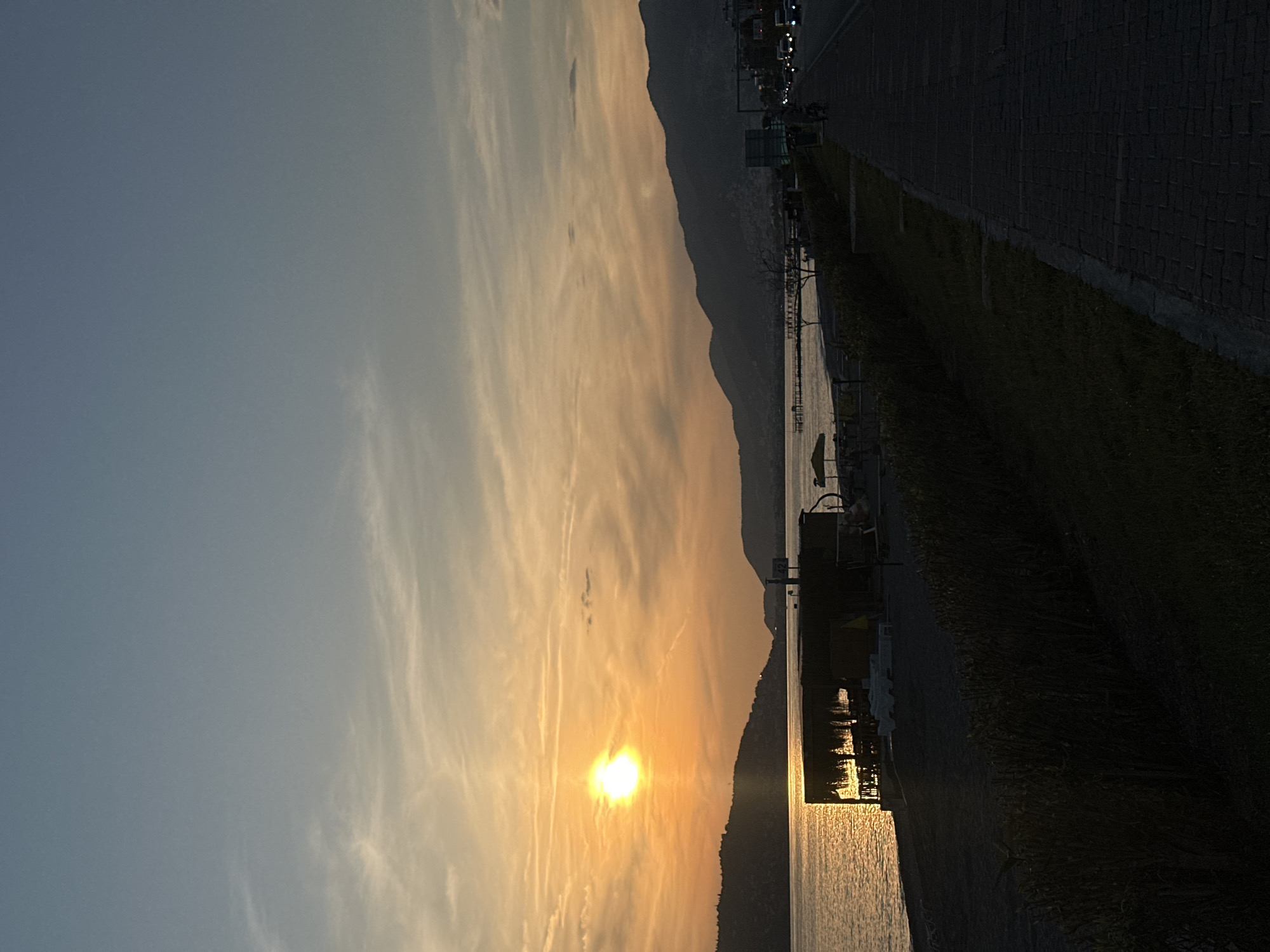 The image captures a sunset over the Mediterranean Sea in Alanya, Turkey. The sun, positioned near the horizon, casts a golden glow across the sky with wispy clouds. A dark, silhouetted mountain range extends along the coastline. A paved walkway runs along the right side of the image, adjacent to a grassy area. Streetlights and vehicles are visible along the walkway. In the sea, the sun's reflection creates a shimmering path of light. Structures, possibly piers or docks, are present along the coastline.
