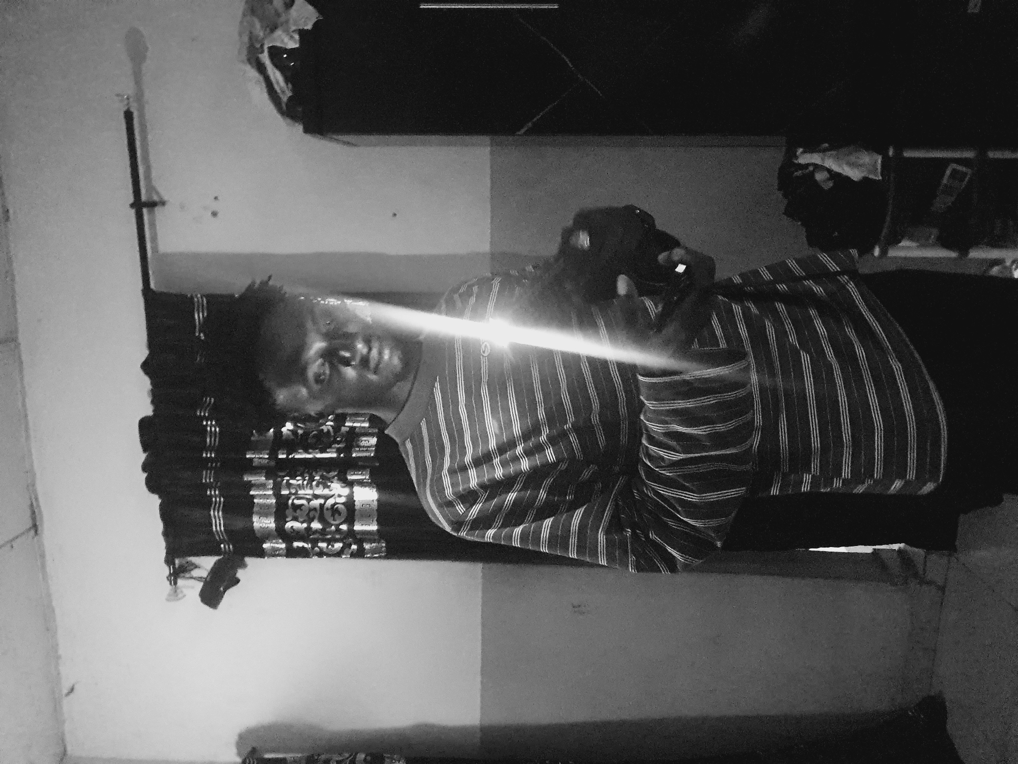 A black and white image depicts a person lying on a bed within a room. The person is facing the camera with their head resting on the headboard. They are wearing a striped shirt. The background shows a wall and other indistinct objects. There is a glare obscuring parts of the image. The location of Lokoja, Nigeria, is not discernible from the visual information provided.