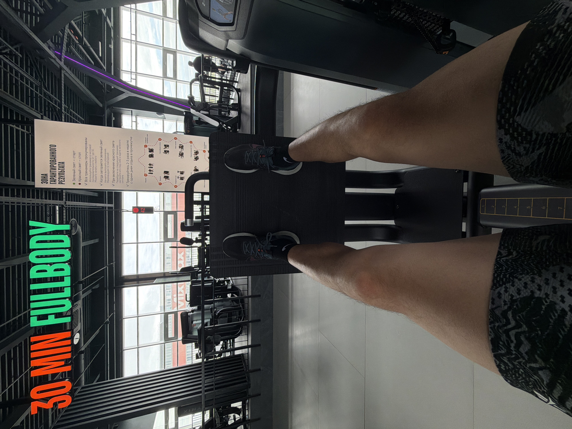 Here is a detailed description of the image:
**Scene:** The image shows a first-person perspective inside a gym. The person is standing on a leg press machine, with their feet on the footplate. They are wearing gray running shoes and patterned shorts.
**Environment:** The gym is modern with large windows offering a view of buildings outside, which suggest an urban setting. A sign on the left wall is written in Russian. The text “30 MIN FULLBODY” is also visible on the left. Exercise equipment is visible around the gym.
**Location:** Based on the sign written in Russian, this gym is located in Russia. The number "42" along with the window view from the gym strongly suggest the city of Krasnoyarsk.
**Objects:** Leg press machine, running shoes, patterned shorts, gym sign, windows, exercise equipment.