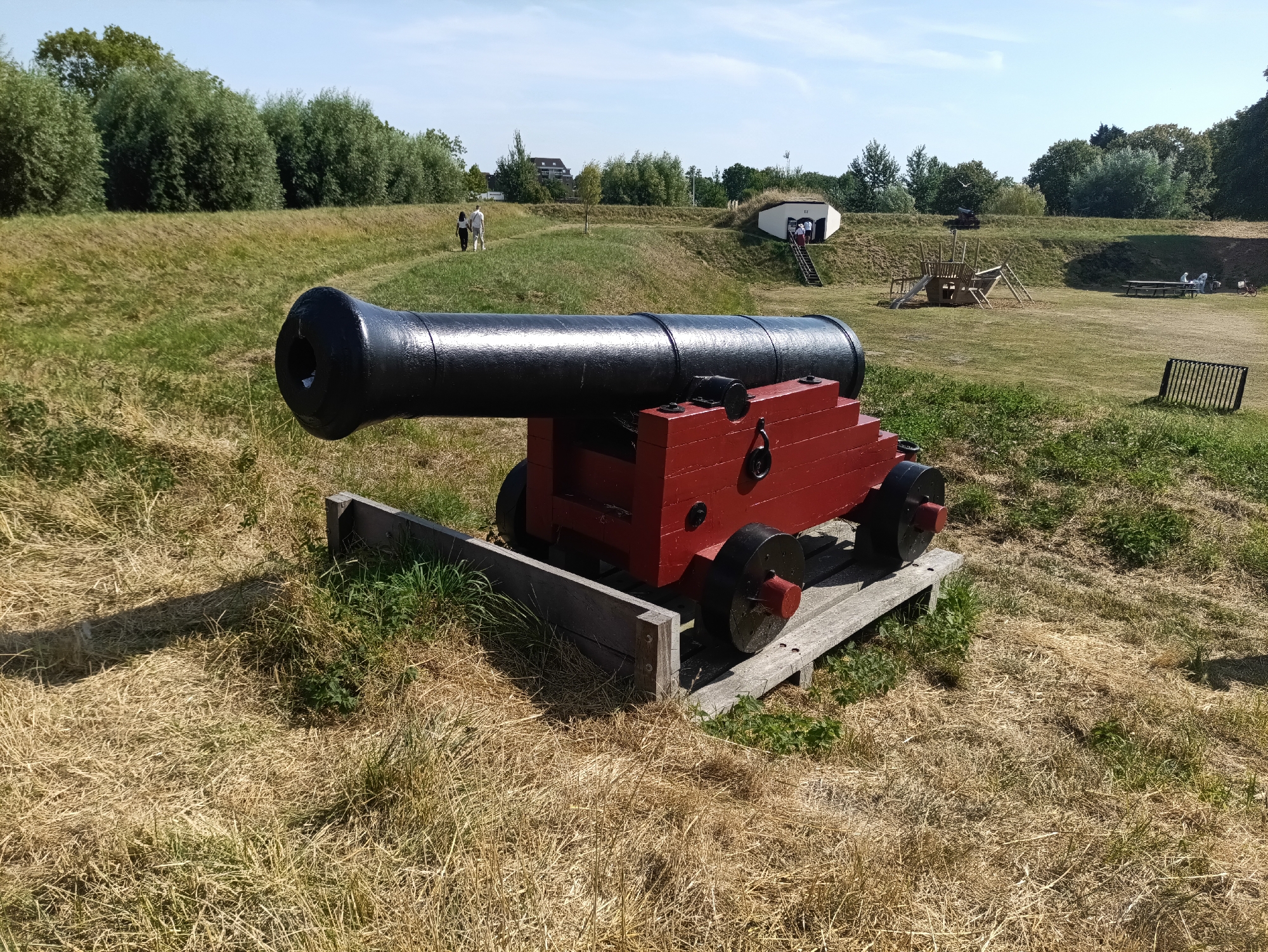 In the city of Brill, Netherlands, a historical cannon is prominently displayed on a wooden platform in a grassy, open area. The cannon is black with a red carriage. Behind the cannon, the landscape consists of rolling green hills dotted with trees. Two individuals, a man and a woman, are walking on a path in the middle ground. Further in the background, a white structure with a green, grass-covered roof is visible, with steps leading up to its entrance. To the right, a wooden playground structure and picnic tables with people are also present. The sky is clear and blue, indicating a sunny day. The foreground is covered with dry, yellowed grass.