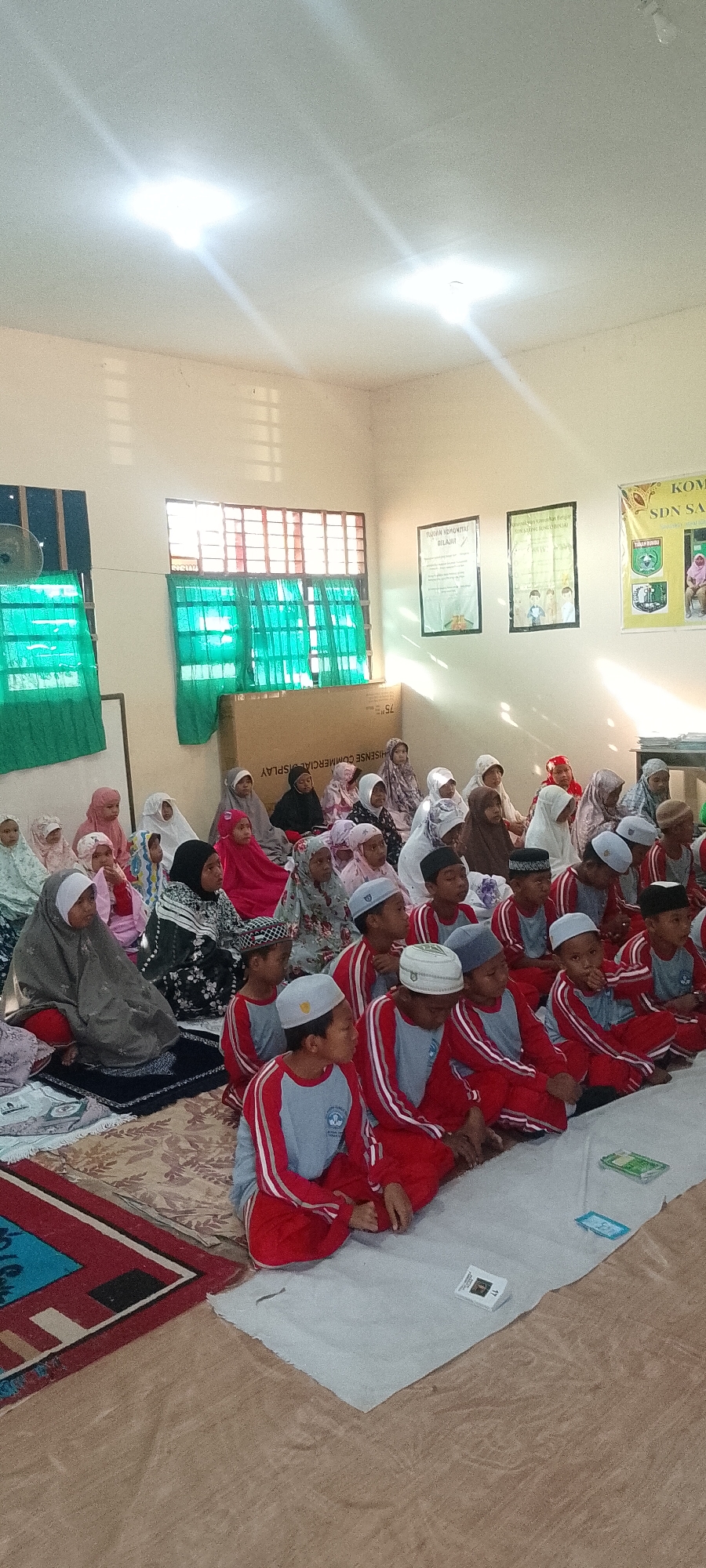 In Saring Sungai Binjai, Indonesia, an indoor scene depicts a group of children, likely students, seated on the floor. They are primarily in two rows, facing the same direction. The children in the front row are wearing red and gray shirts with white stripes on the sleeves, and some have on caps. The children in the back row are wearing a variety of hijabs and outfits. The room appears to be a classroom or communal space, with large windows covered by green curtains. A large cardboard box, labeled "HISENSE COMMERCIAL DISPLAY", is situated behind some of the children. Two framed posters are hung on the wall behind the seated children, one of which is partially visible and appears to be a school banner for "SDN SA...". The lighting suggests an interior space, with bright ceiling lights casting illumination. The floor is covered with rugs and mats. Some of the children are looking forward, as if attending to a speaker or activity.