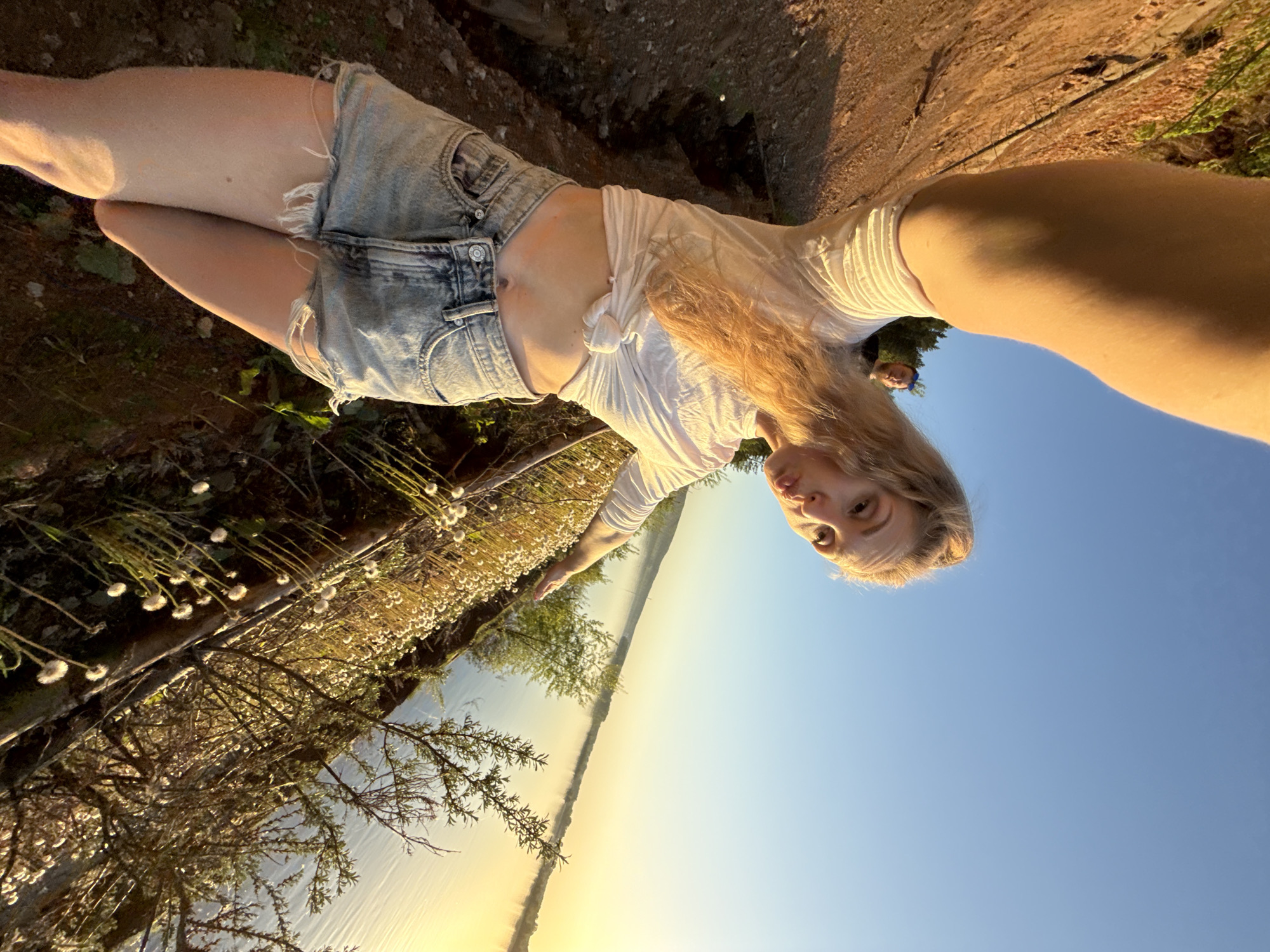 The image depicts a woman in denim shorts and a white t-shirt standing outside near a body of water under a clear, blue sky. She appears to be on a slightly elevated area with vegetation nearby, including trees and small white-flowered plants. The water stretches towards the horizon with a golden glow. It could be the Kama River in Chaikovsky, Russia, due to its wide expanse and location near tree-lined areas.