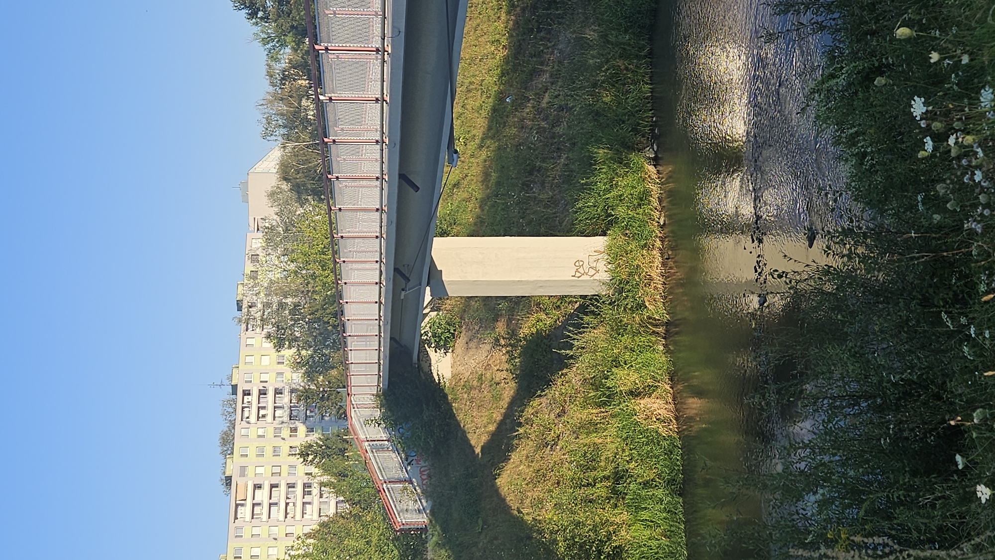 A pedestrian bridge spans across a river in Prešov, Slovakia. The bridge, made of concrete and metal, is supported by a single concrete pillar in the middle of the riverbank. The sides of the bridge are lined with a metal railing. Green grass and trees surround the bridge, with an apartment building visible in the background. The river flows calmly, reflecting the sky and surrounding foliage. There is graffiti on the concrete pillar of the bridge. The sky is clear and blue, indicating a sunny day.