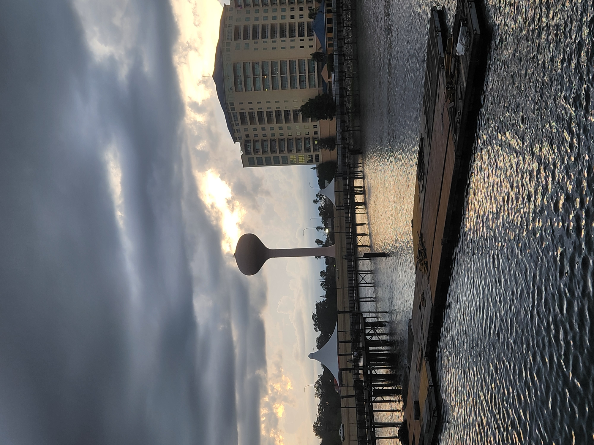 This image captures a waterfront scene in Altamonte Springs, Florida, United States of America, during a cloudy sunset. In the background, a tall, light-pink water tower stands prominently against the dark, dramatic sky. To its right, a multi-story beige building with numerous balconies and windows is visible. In the mid-ground, a wooden pier extends over the water, with a white-canopied structure and some people standing on it. The water in the foreground is rippled, reflecting the muted light of the setting sun. Two dark-colored barges or floating docks are partially submerged in the water in the immediate foreground. The overall atmosphere is somber due to the overcast sky, with breaks of light suggesting the sun is attempting to break through the clouds.