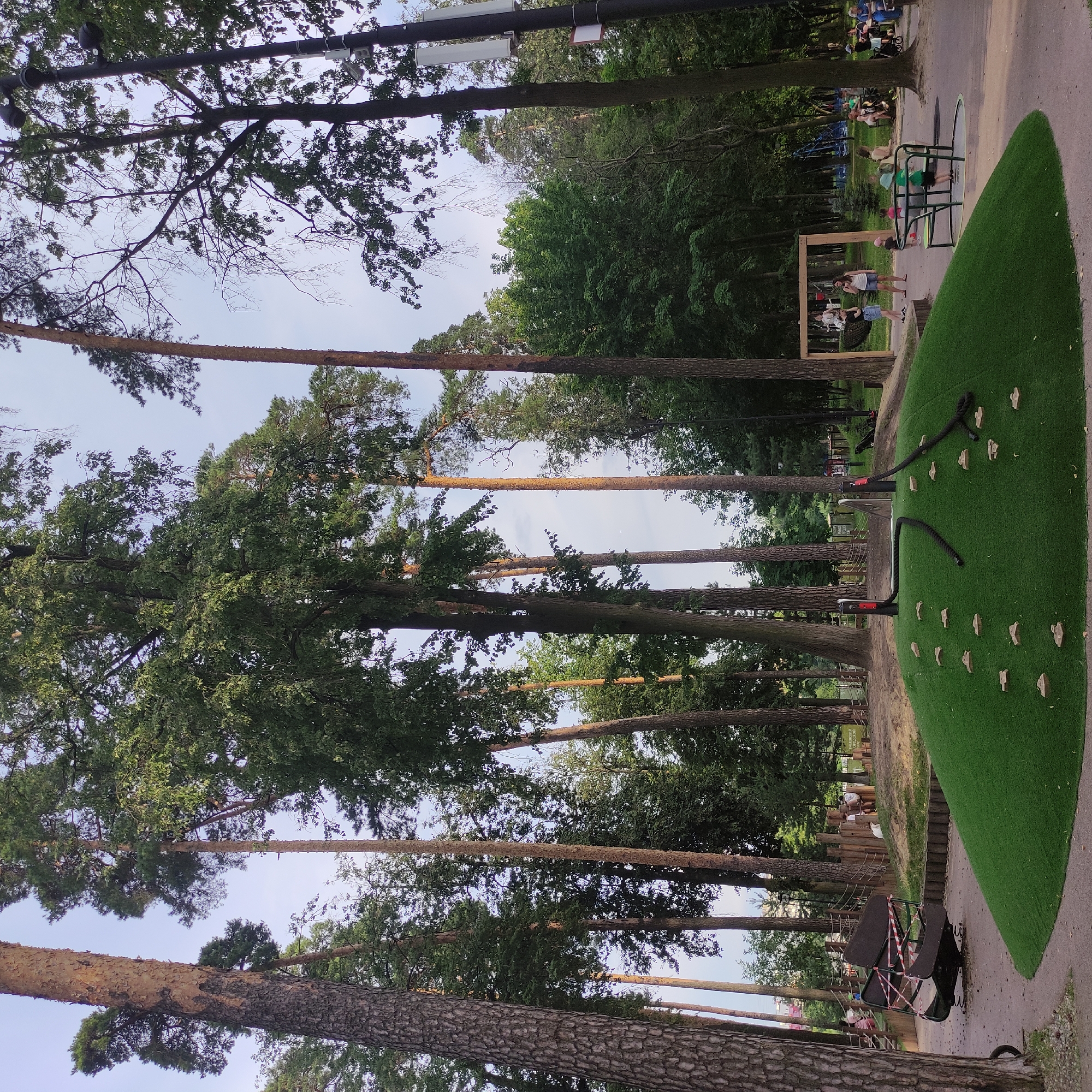 This image depicts a park in Ramenskoye, Russia, featuring a playground area and surrounding trees. In the foreground, a green, grassy mound with climbing holds and ropes is visible. To the left, a broken playground ride, cordoned off with red and white tape, is situated on a paved surface. Further back, several people are present, including children playing on a merry-go-round and adults sitting on benches or walking. A wooden swing set is also visible. The background consists of tall trees, characteristic of a forested park environment, with a partly cloudy sky overhead. The scene appears to be a public recreational space.
