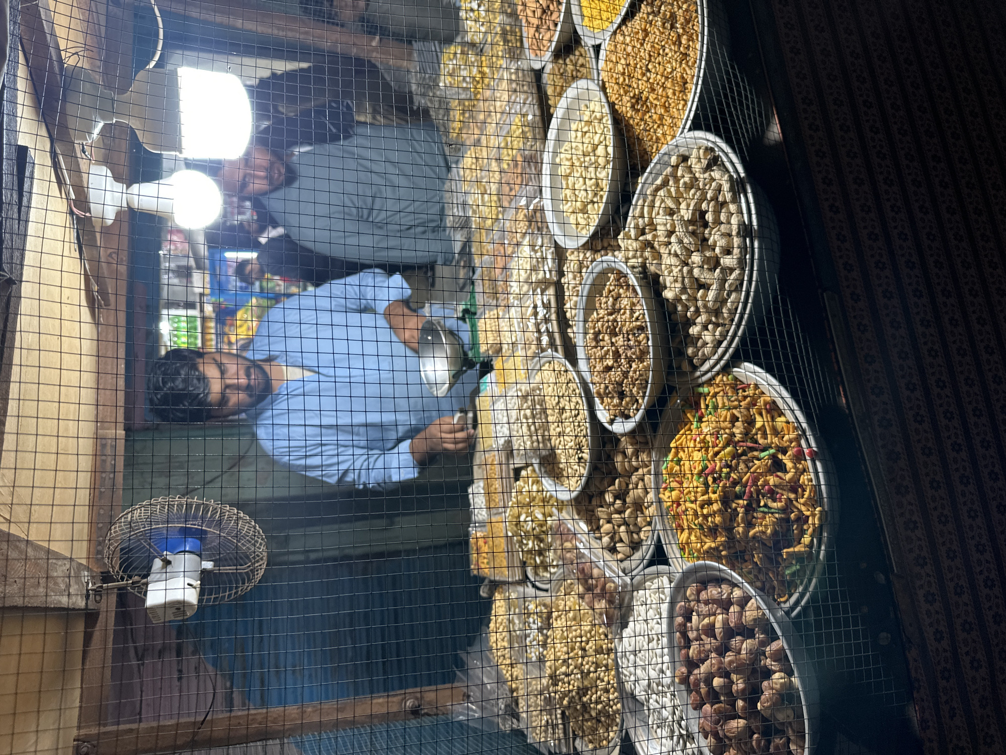 This image captures a vibrant snack stall, viewed through a prominent wire mesh enclosure, likely at a market during the evening or night. The stall features a wide array of dry fruits and savory snacks displayed in numerous round and oval metal trays. Visible items include peanuts in their shells, shelled nuts like almonds and walnuts, dried dates, and a colorful, crunchy savory mix often found in South Asian markets.
Two men are visible within the stall. The man in the foreground, wearing a light blue shirt, appears to be the vendor, standing behind the displayed goods and looking towards the right side of the frame. Another man in a grey shirt is seen further back, partially obscured. The stall is brightly illuminated by several exposed LED lightbulbs, casting strong light on the food and the men. A small, wall-mounted electric fan is also visible on the wooden structure of the cage. The wire mesh creates a distinct pattern across the entire image, separating the viewer from the vendor and his products. The background behind the food display is dark with a subtle patterned fabric. The overall scene suggests a bustling, utilitarian market environment, characteristic of street vendors or small shops in regions like South Asia or the Middle East, where such snacks are popular. There is no visible text in the image.