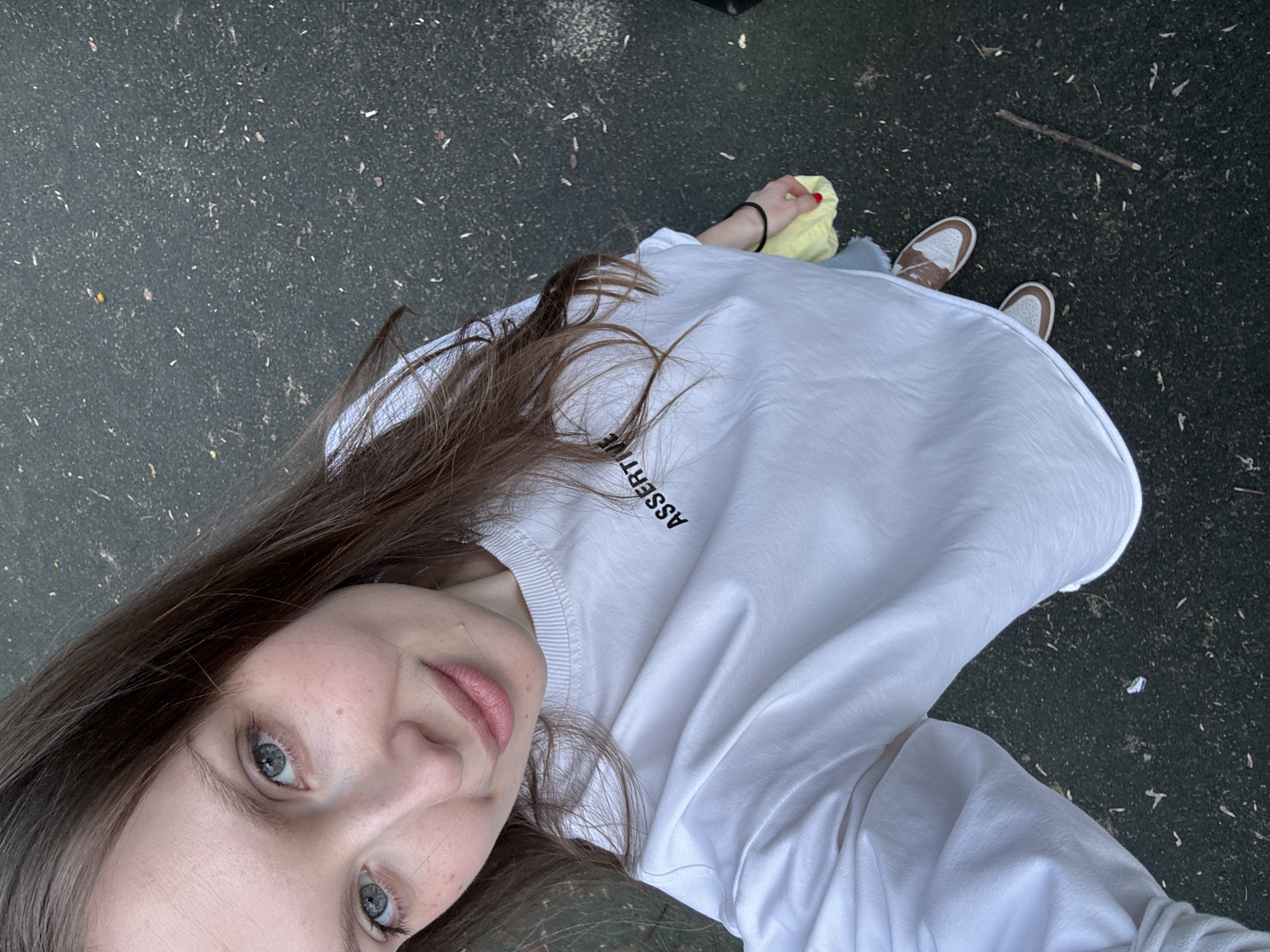 A woman with long brown hair, wearing a white sweatshirt with the word "Assertive" printed on the upper left chest, is taking a selfie from a high angle, looking up at the camera. She has fair skin and light-colored eyes. In her hand, she is holding a yellow cloth. She is standing on asphalt that appears to be outdoors, with a stick nearby. Her shoes are visible, a pair of white sneakers with brown accents. The location is not identifiable as Moscow, Russia, from the visual information provided.