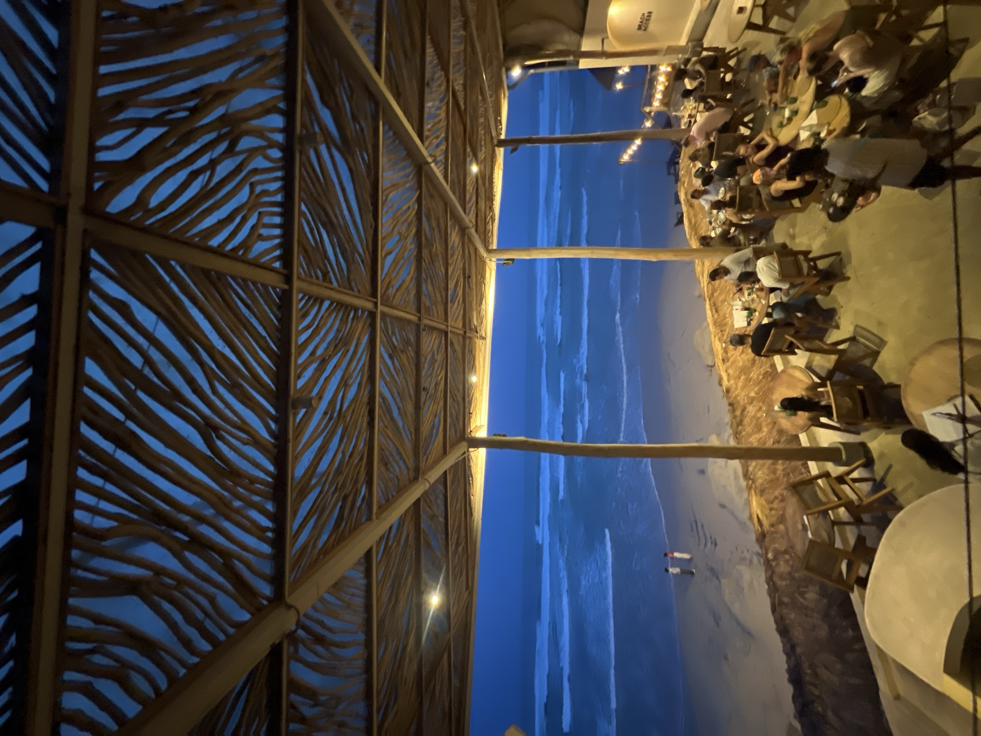 The image shows an outdoor dining area at a beachside restaurant in Ahangama, Sri Lanka. The view is oriented with the beach and ocean stretching horizontally across the middle of the frame. A decorative wooden structure, possibly part of a roof or awning, occupies the left side, with sunlight filtering through its gaps. On the right, people are seated at tables and chairs, suggesting a dining or social activity. Some individuals are identifiable, but the details of their clothing or actions are somewhat blurred. The background shows the beach transitioning into the ocean, which appears vast and dark blue. A light source illuminates the dining area, with additional lights visible further away. The image is taken at an angle, presenting a perspective looking out from the restaurant towards the beach.