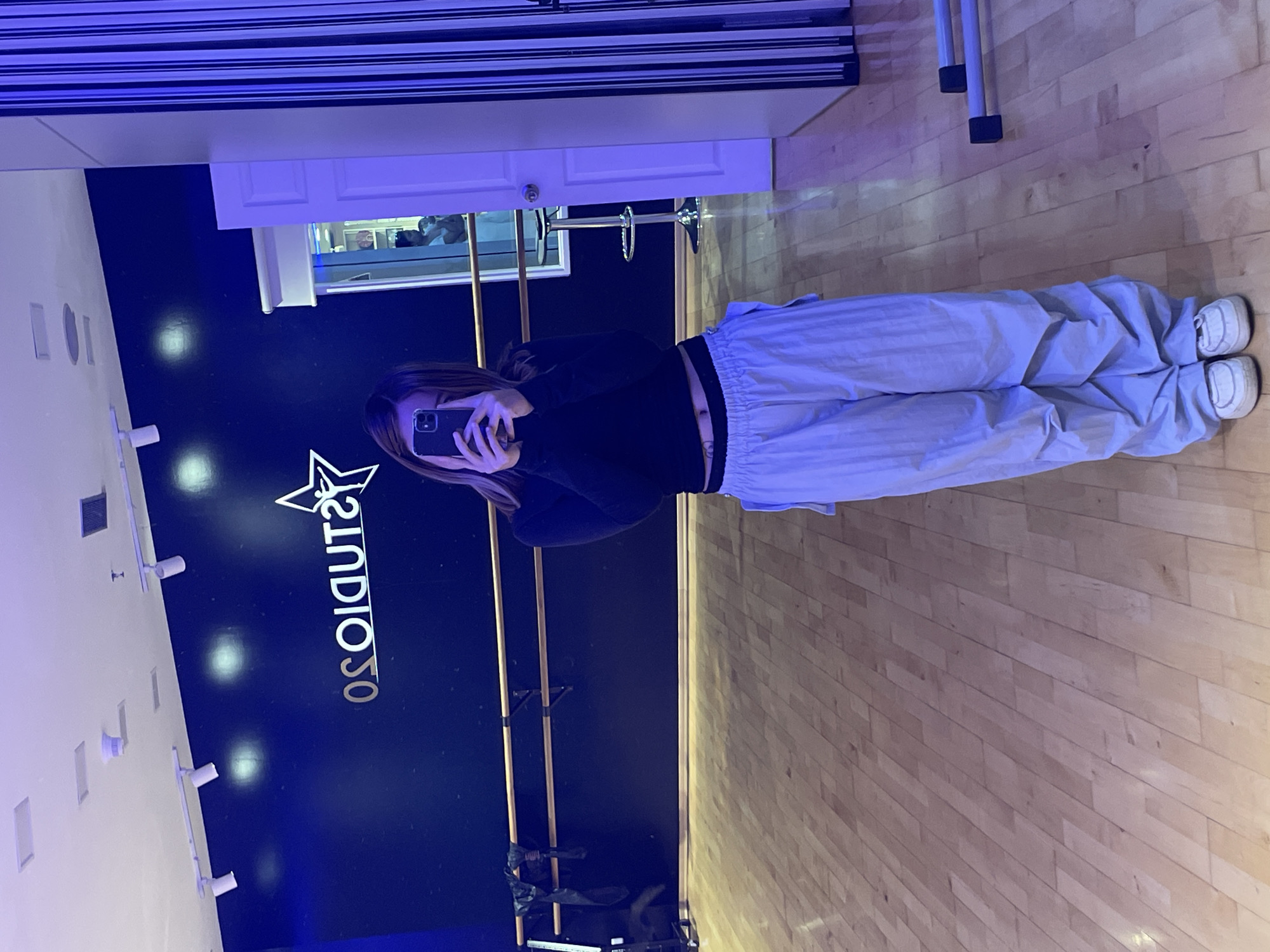 A person with long brown hair is taking a picture of themselves in a mirror. The person is wearing a black sweater and light blue sweatpants, showing a bit of their stomach. The location is likely a dance studio, suggested by the "Studio Solo" signage and ballet barres. The background is predominantly a dark blue wall with the studio name and star logo. The floor is light wood, and there is a white shelf with items on it above the mirror. The reflection in the mirror also shows what appears to be a doorway or window with a bright light coming through. The picture gives no indication of the city of Porlamar, Venezuela.
