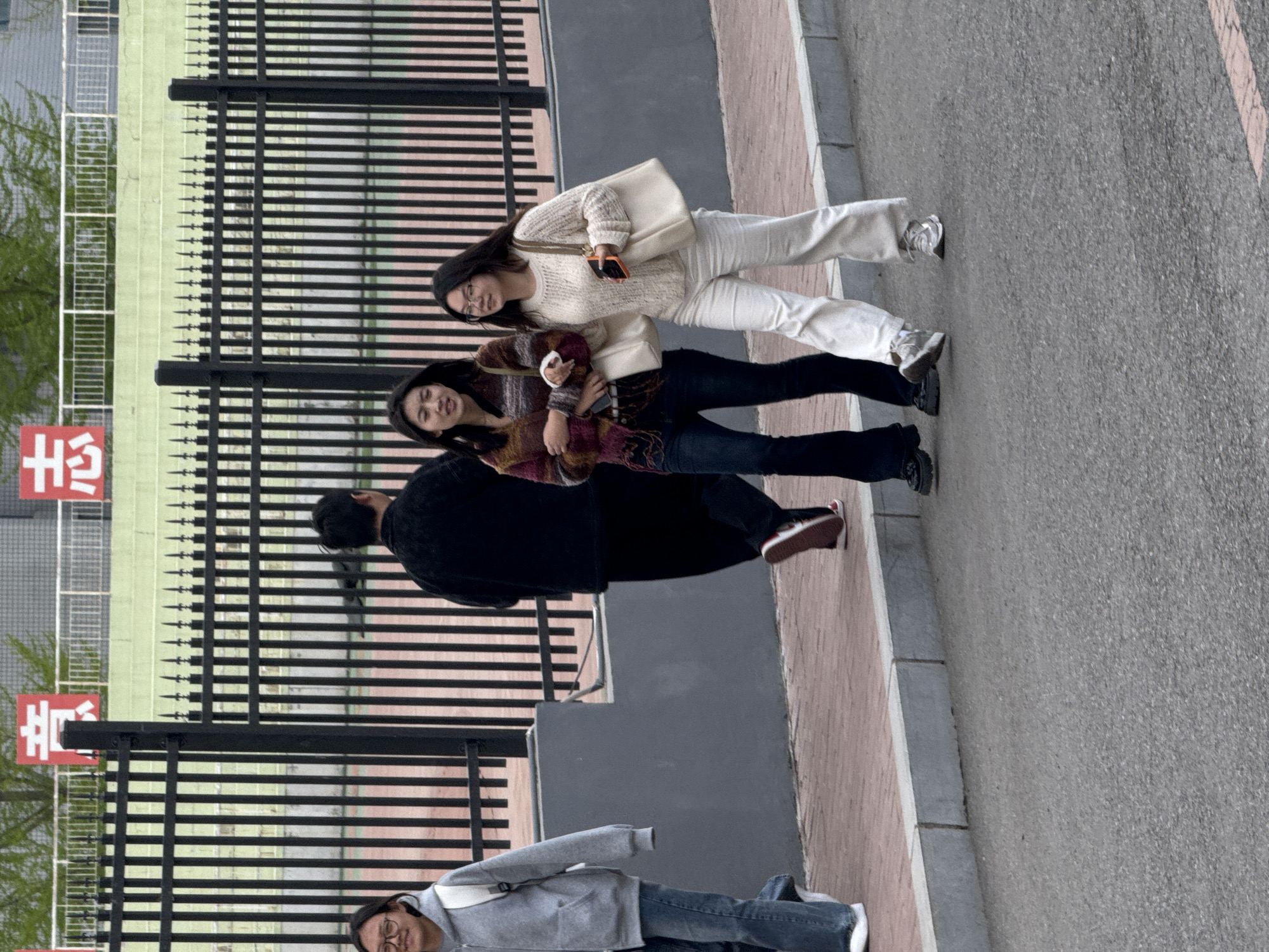The image captures an outdoor scene, likely in an urban or campus environment in Zhongtun, China, featuring four individuals on a paved sidewalk next to an asphalt road. A tall, black metal fence with horizontal bars and pointed vertical elements separates the sidewalk from a light green wall or building and some foliage in the background.
In the foreground, four people are visible. On the right, two young women walk together. The first woman wears a cream-colored knitted sweater, white wide-leg pants, and white sneakers, carrying a light-colored shoulder bag and holding a mobile phone. She has long dark hair and wears glasses. Slightly behind her, the second woman wears a multi-colored (brown, burgundy, cream) striped sweater, dark blue jeans, and dark shoes. She also has long dark hair.
Behind these two, partially obscured by the fence, a third person is seen from the back, wearing a black top and dark pants, with brown sneakers. Further to the left and slightly lower on the sidewalk, a fourth person, also with long dark hair and glasses, wears a grey hoodie, blue jeans, and white sneakers, looking directly towards the camera.
The sidewalk is made of light-colored, possibly reddish-brown, pavers, bordered by a grey curb leading to the grey asphalt road. The time of day appears to be daytime, possibly overcast, given the lack of strong shadows. A red sign with white Chinese characters is visible behind the fence; the top character clearly reads "出" (chū), meaning "exit". The bottom character is partially obscured.