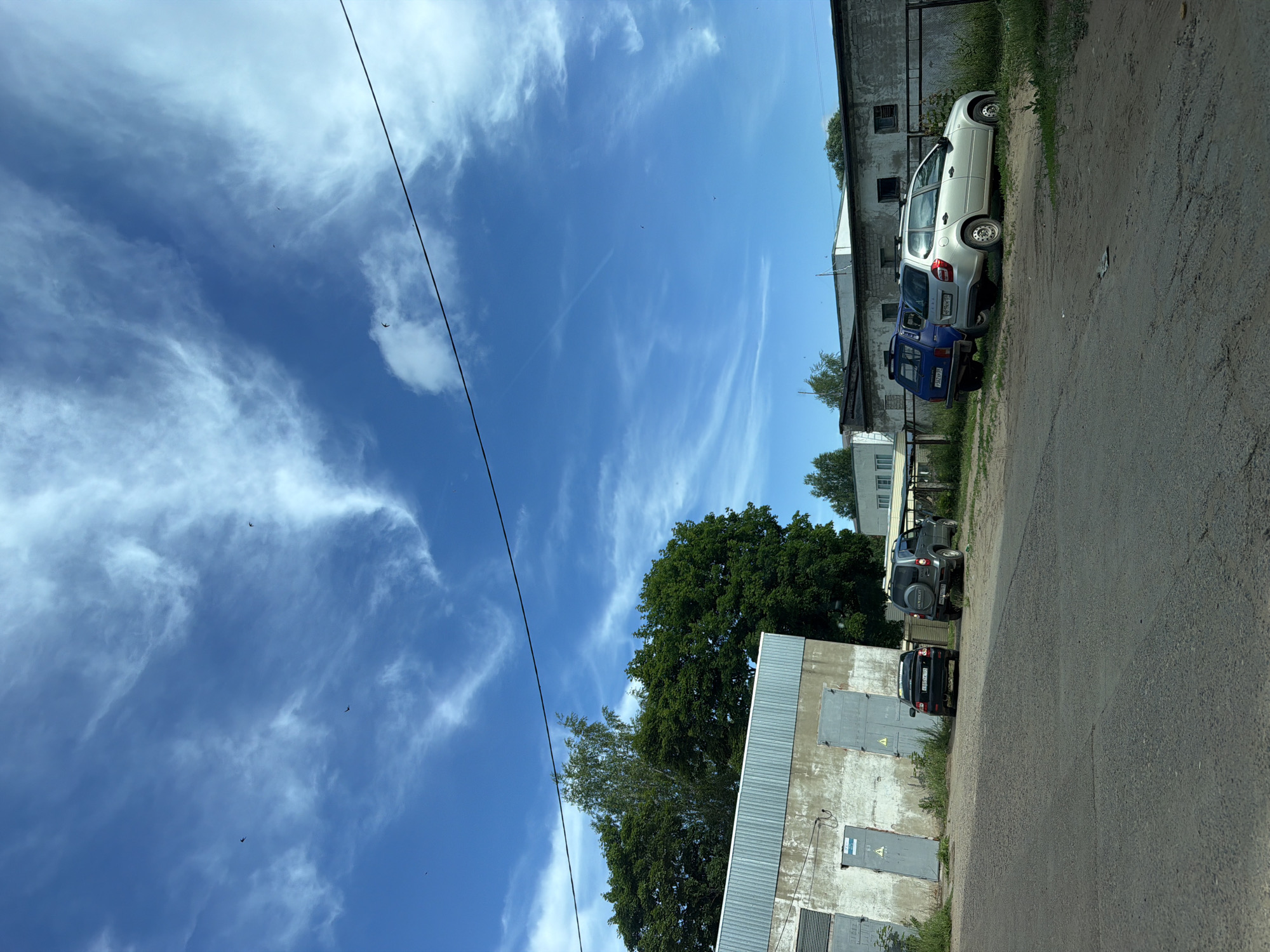 The image depicts an outdoor scene with several vehicles parked in front of industrial-style buildings under a bright blue sky with wispy clouds and several small birds in flight. On the left, a dark-colored sedan is parked next to a gray SUV. Further to the right, a blue minivan and a silver station wagon are parked. The buildings appear to be warehouses or garages, constructed with concrete and corrugated metal. Lush green trees are visible behind the buildings on the left side of the image. The ground in the foreground is a rough asphalt or dirt surface. The scene suggests an industrial or commercial area, possibly within the city of Chaikovsky, Russia, given the context.