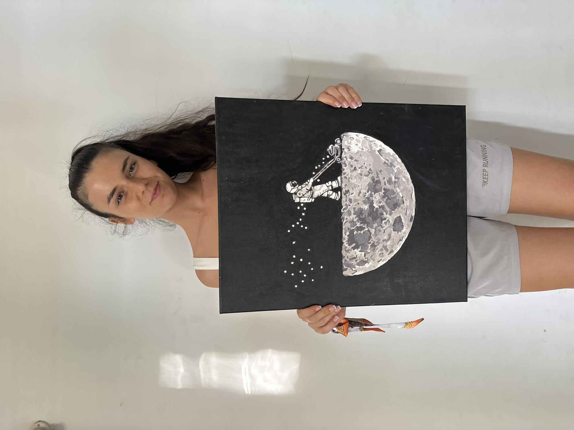 A woman is holding up a black square canvas with a painting on it. The painting depicts a moon, partially visible on the right side of the canvas, with an astronaut scaling it. The astronaut appears to be breaking off small white pieces from the moon, which are depicted as scattered white dots below. The woman has long, dark hair and is wearing a white tank top and gray shorts with the words "KEEP RUNNING" printed on the side. She is smiling and looking at the camera. The background is a plain white wall. The location is not discernible from the image.