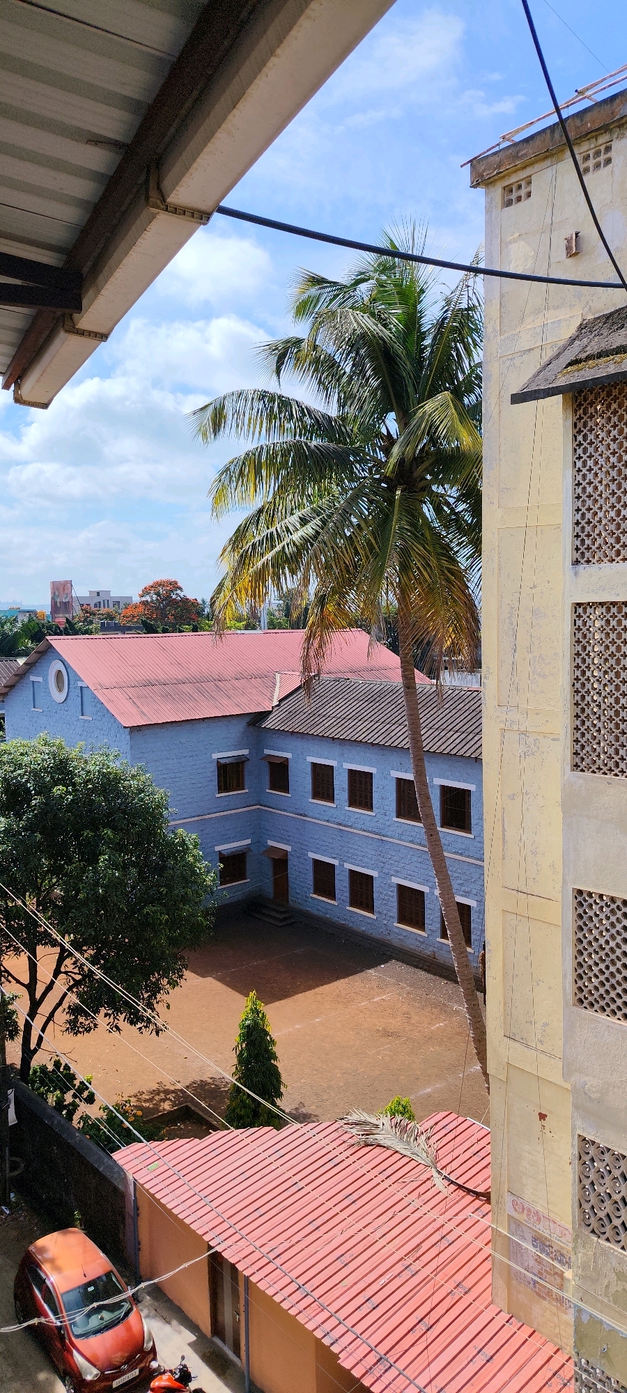 A high-angle, vertical, medium-shot captures a scene on a partly cloudy day in Belagavi, India, showcasing a courtyard and adjacent buildings. A tall, slender palm tree, its fronds partially obscuring the sky, dominates the center of the frame. To the left, a two-story, light blue building with a red corrugated metal roof is visible. It features several brown-paned windows and a circular decorative element on its upper facade. A large, leafy green tree stands to the left of this blue building. In the foreground, a small, orange car is parked on a red tiled roof next to a dark wall. A motorcycle is partially visible behind the car. To the right, a section of a tall, pale yellow building with decorative concrete screens is present. The ground in the courtyard appears to be dirt. The overall environment suggests a residential or institutional setting within a city.