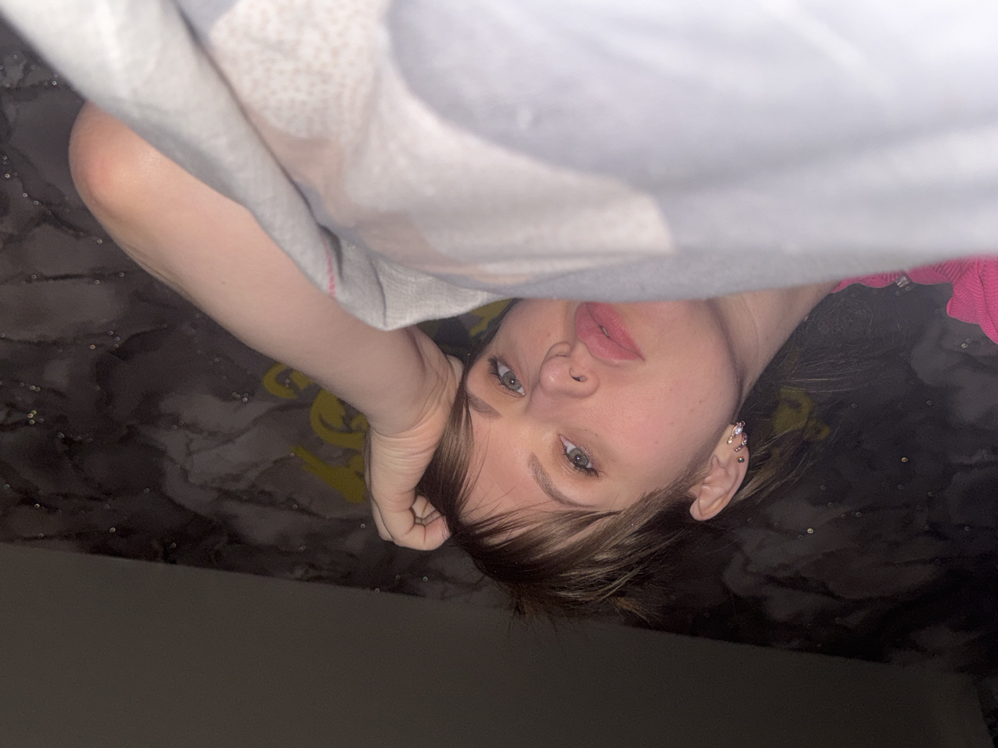 This rotated, close-up portrait features a person, likely female, lying down and looking directly at the camera, appearing upside down. A light-colored, possibly grey or white, blanket or fabric covers the upper half of the image, partially obscuring the person's forehead and upper body.
The subject has light skin, piercing blue-grey eyes, and medium-brown hair with bangs. Their lips are a soft pink. One hand is visible, resting casually near their head. On their visible right ear, several decorative piercings are apparent, including tragus, helix, and lobe piercings with small, shiny earrings.
A hint of a pink or magenta garment is visible on the right side of the frame. The background behind the person's head is a dark, possibly black or dark grey, surface featuring a subtle swirling pattern and scattered tiny reflective specks. A faint yellow shape or design can be seen in the upper-left background. The lower edge of the image is very dark. The dim, focused lighting on the face suggests an indoor setting, possibly during evening hours. There is no visible text. The image conveys a casual and intimate mood.
