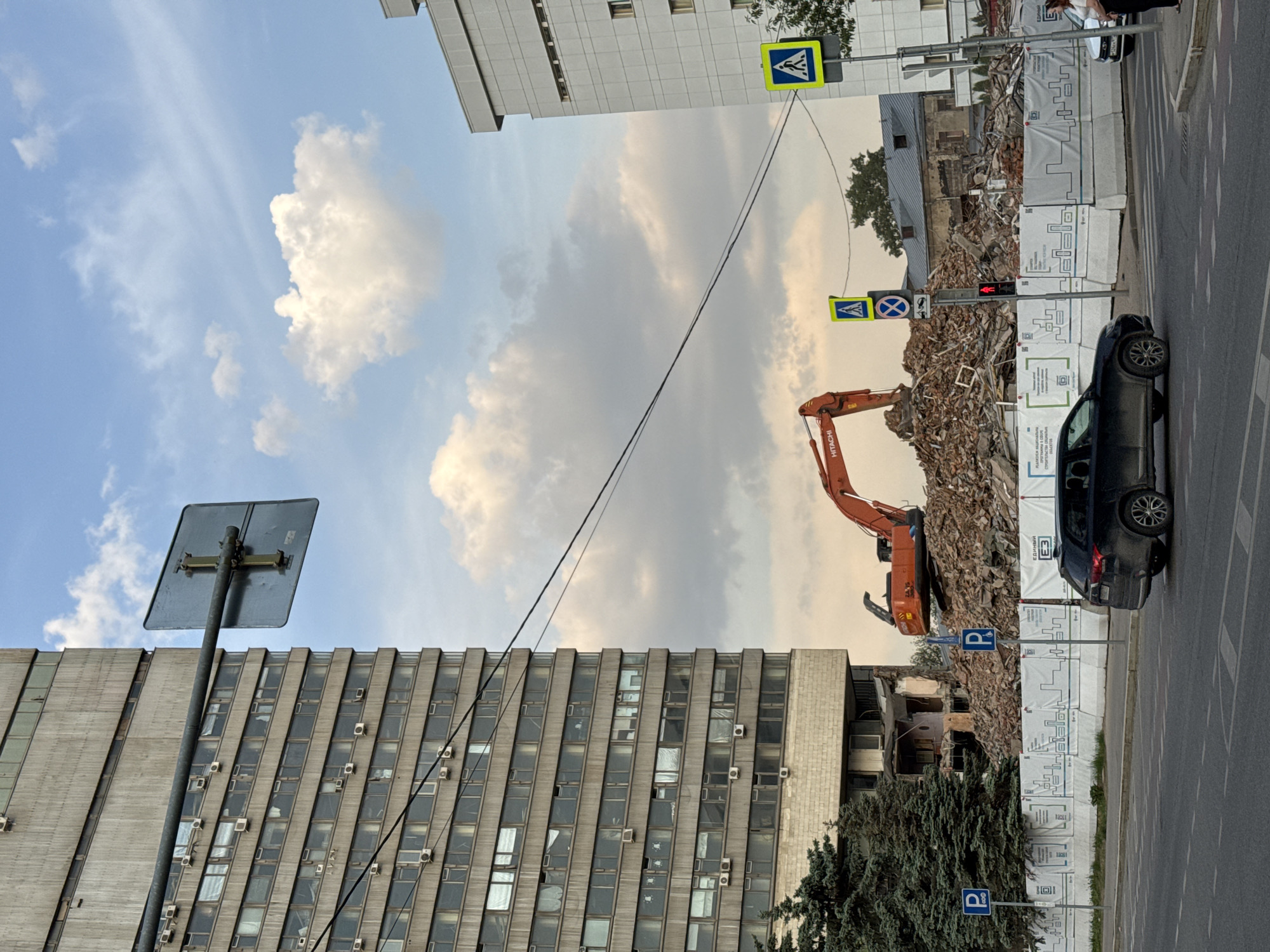 The image depicts a scene of demolition in Moscow, Russia. An orange Hitachi excavator operates atop a pile of rubble, likely the remnants of a demolished building. The site is partially bordered by white construction barriers displaying repeating patterns. To the right, a black car is parked on the street.
In the background, a multi-story building with a grid pattern of windows and air conditioning units is visible. A power line stretches across the sky. Above the rubble pile, a pedestrian crosswalk sign hangs above the street. Traffic signal on the right-hand side is in the red state.
The sky contains scattered white clouds.