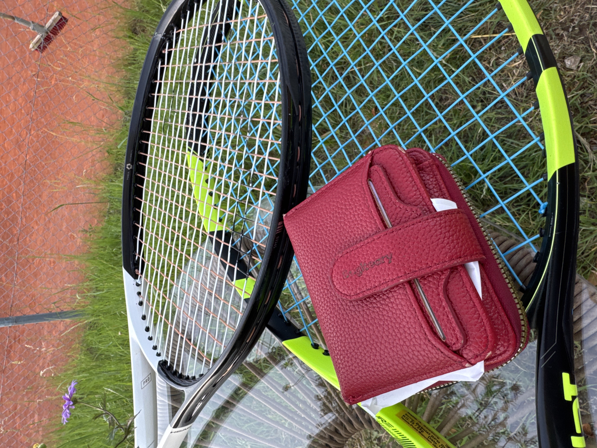 In Neubiberg, Germany, a red leather wallet with embossed lettering lies on top of a tennis racket with a black frame and blue strings. The tennis racket is resting on a glass table, which is set against a background of green grass and a terracotta-colored tennis court. In the upper left corner, a small purple flower blooms amongst the grass. A broom with a wooden handle and red bristles is attached to a chain-link fence in the upper right portion of the image.